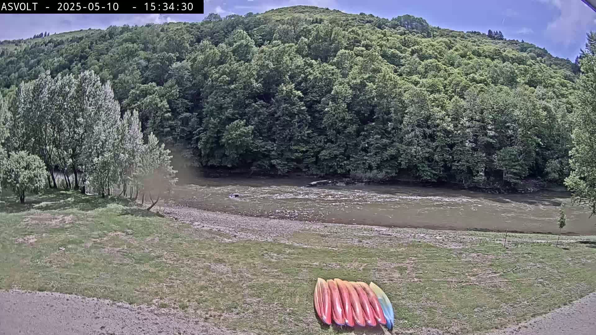 A river flows under a partly sunny sky past a grassy bank where several kayaks are lined up, and lush green trees cover the hills in the background.