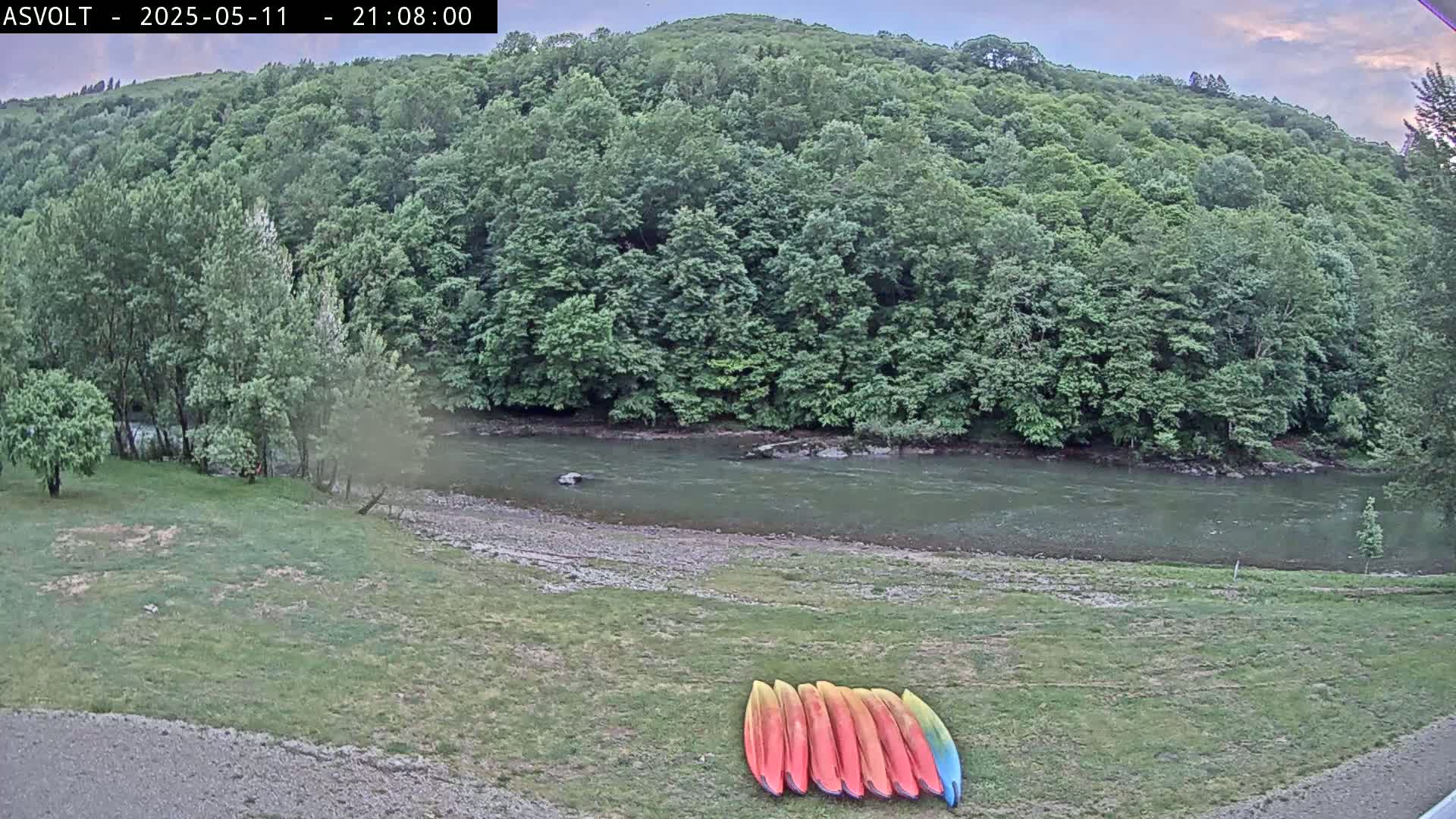 Several kayaks are lined up on a grassy bank next to a calm river under a partly cloudy evening sky, bordered by lush green trees.
