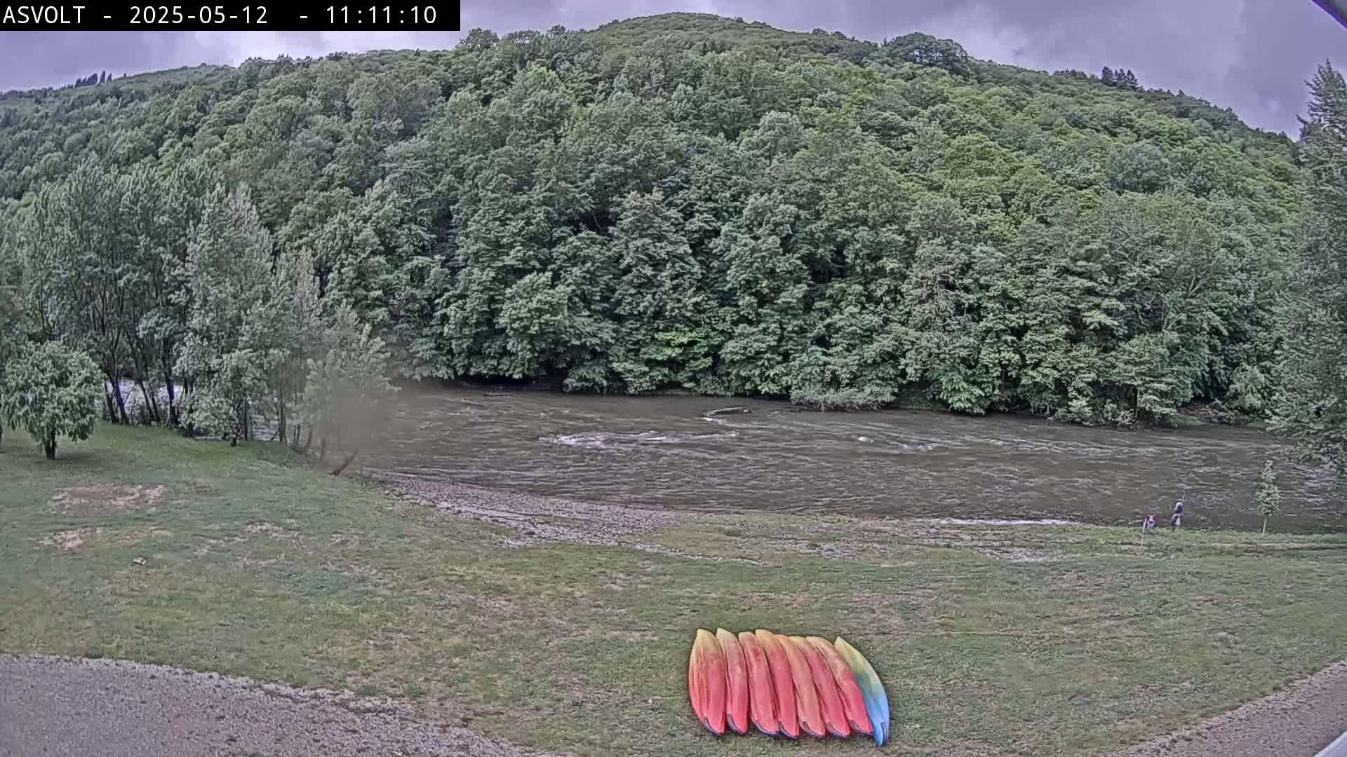A river flows under an overcast sky past a lush green hillside where several kayaks are lined up on the grassy bank.