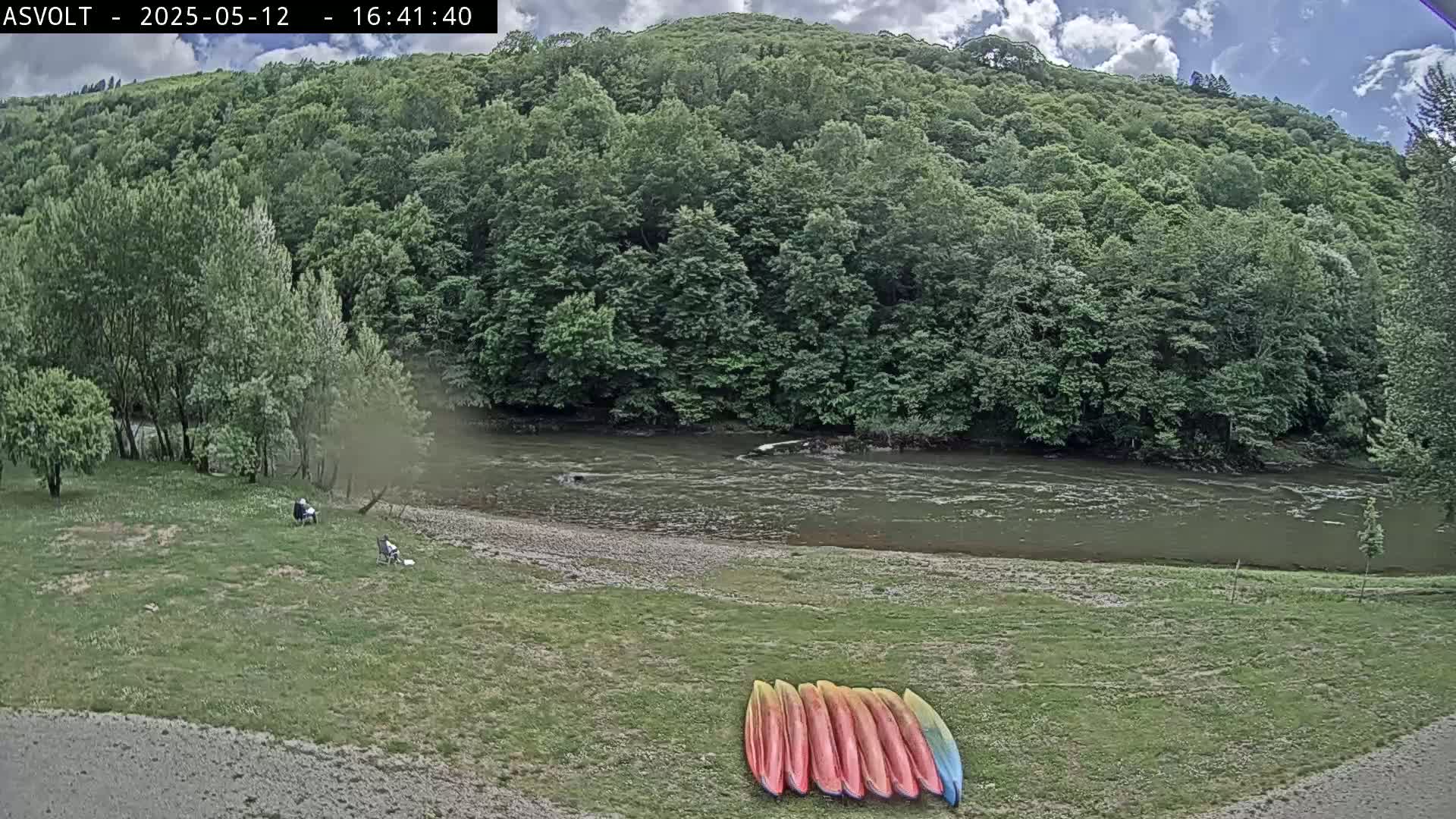 A partly cloudy day shows a river flowing past a grassy bank where several kayaks are lined up, with a forested hillside beyond.