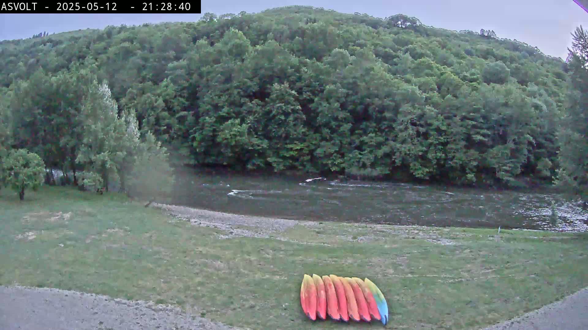 A calm river flows past a grassy bank where several kayaks are stored, surrounded by lush green trees under an overcast sky.