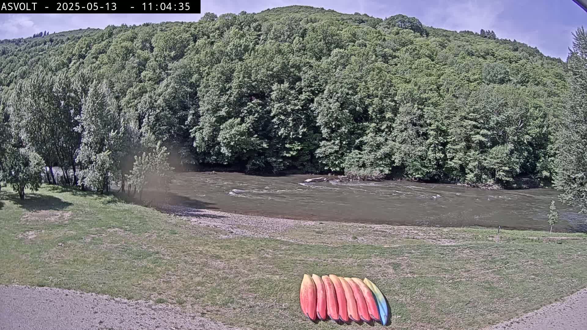 A river flows under a partly sunny sky past a grassy bank where several kayaks are lined up.