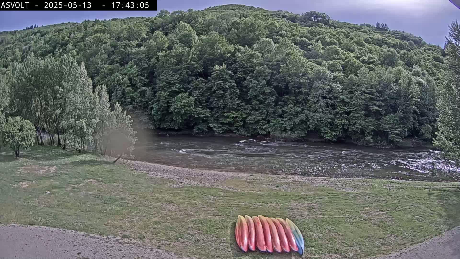 Several kayaks lie on a grassy area next to a flowing river bordered by lush green trees under an overcast sky.