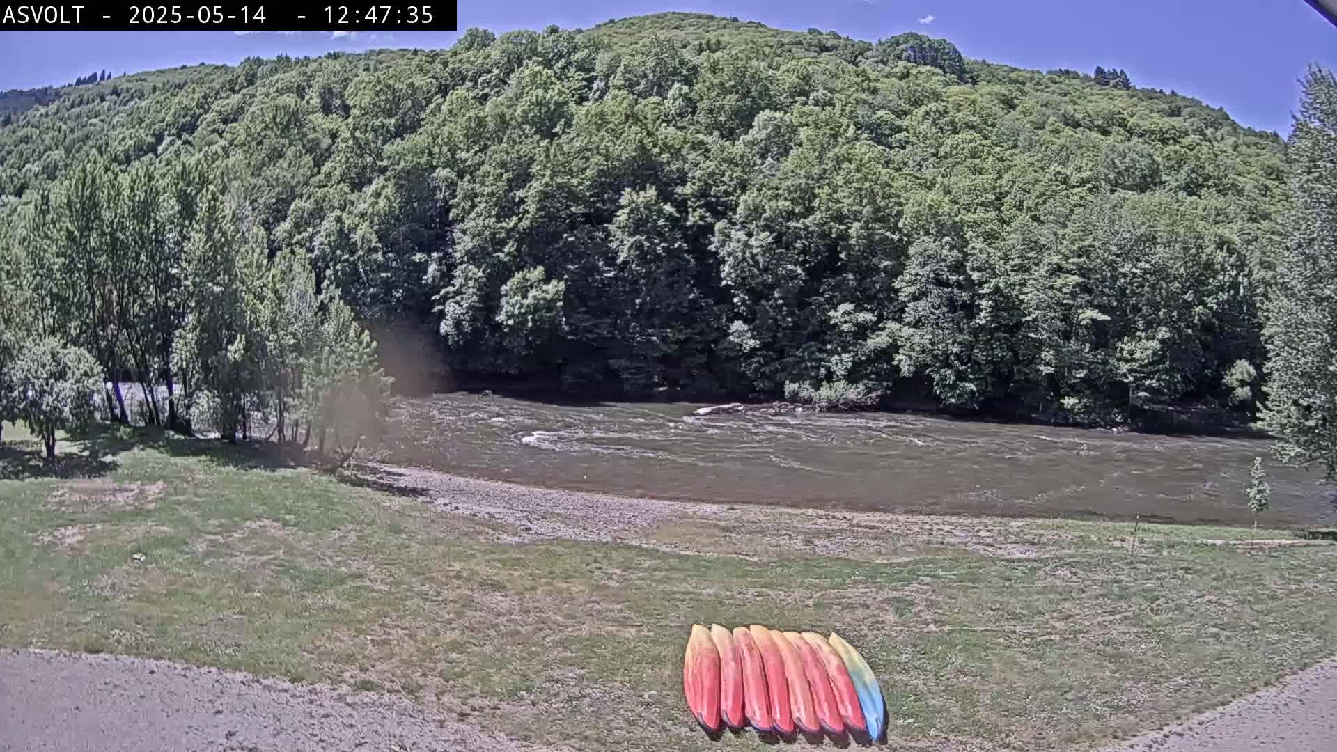 A river flows under a sunny sky past a grassy bank where several kayaks are lined up.