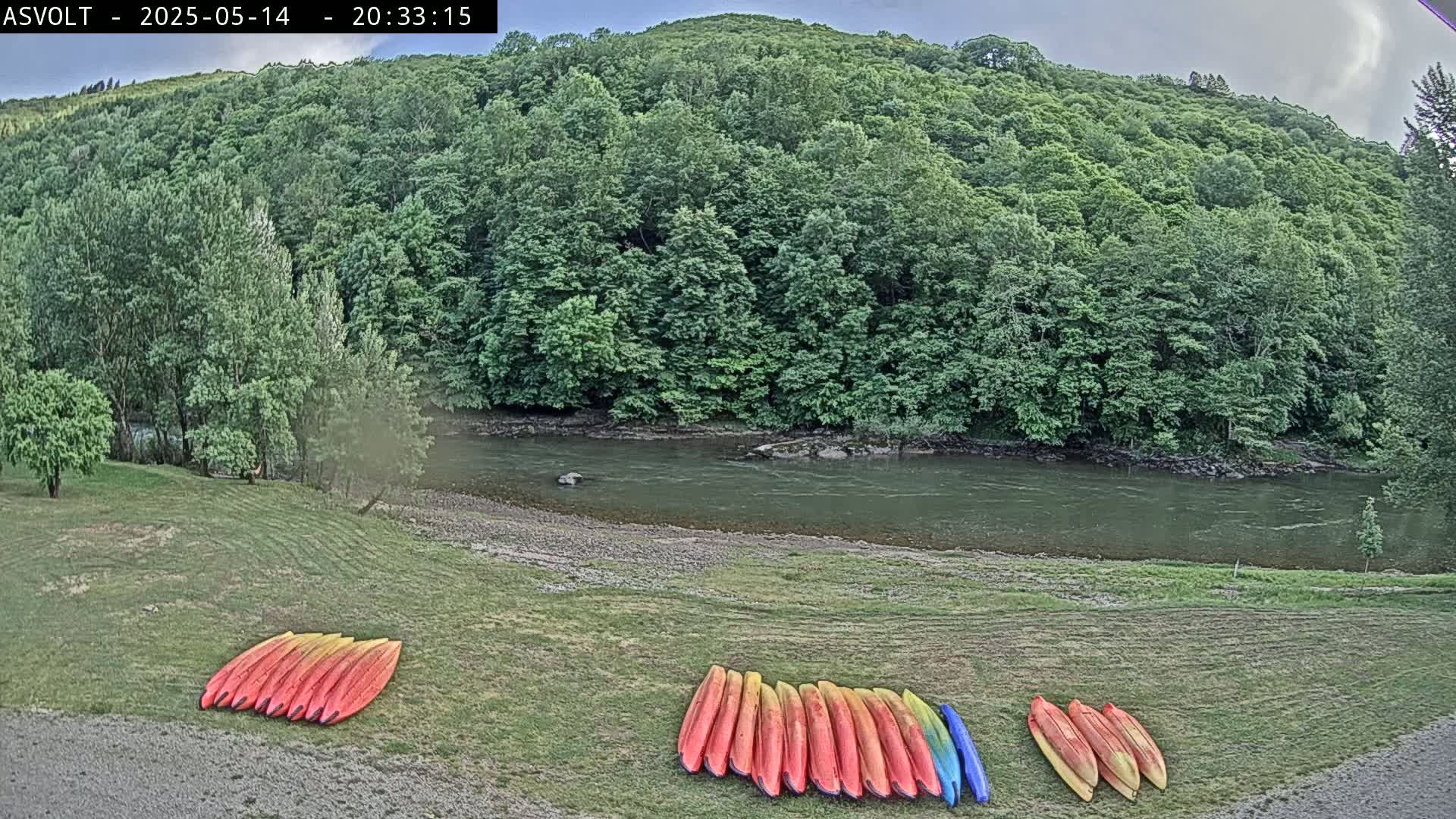 Several kayaks are lined up on a grassy area next to a calm river, with lush green hills and trees in the background under a partly cloudy sky.