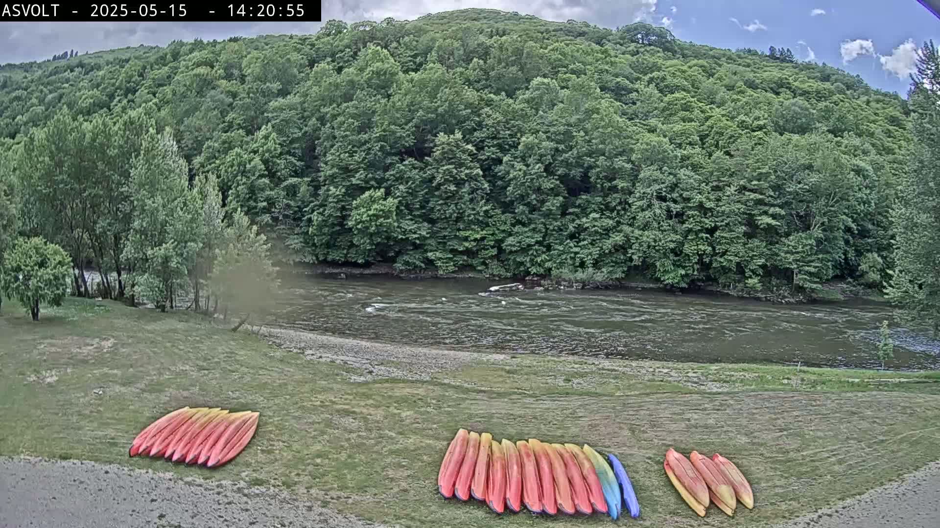 A calm river flows past a lush green forest under a partly cloudy sky, with several kayaks stacked on a grassy riverbank.