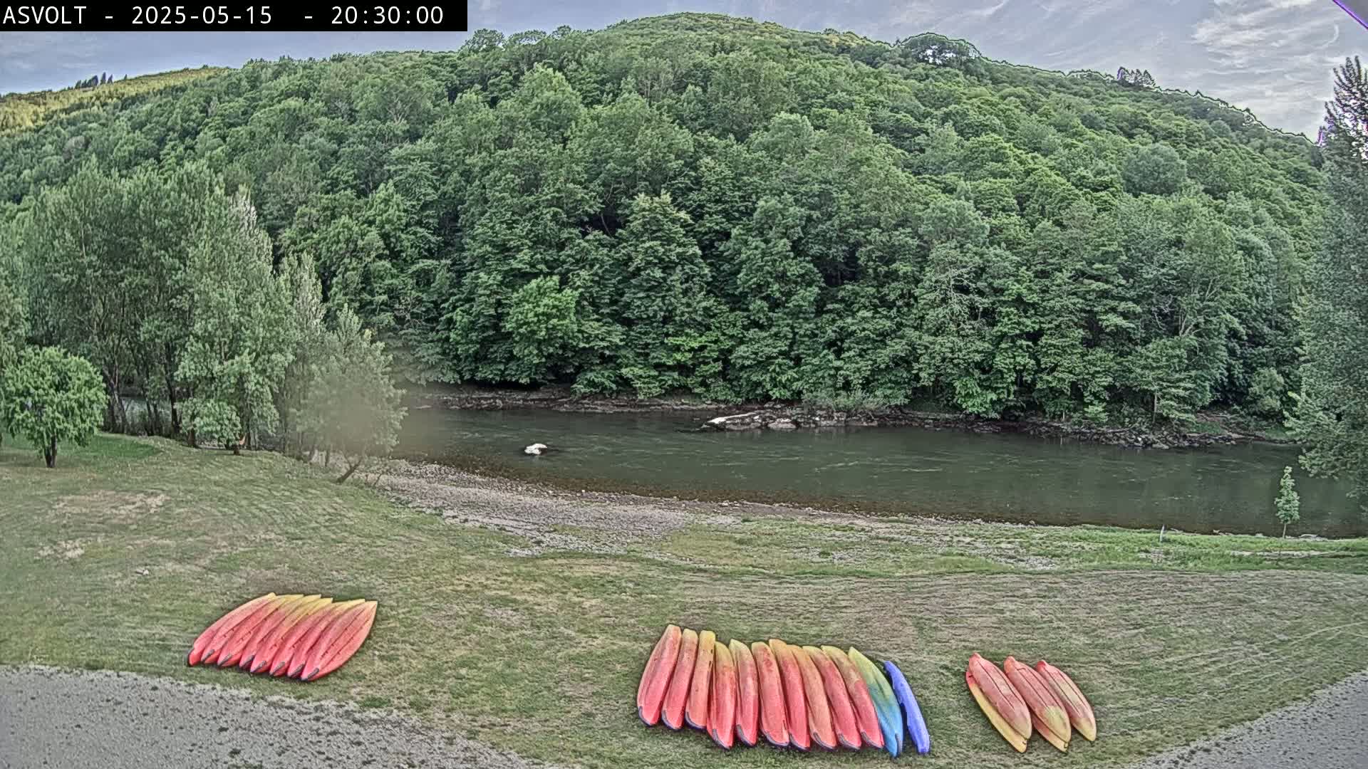 Kayaks are stacked on a grassy area next to a calm river, which is bordered by lush green trees under an overcast sky.