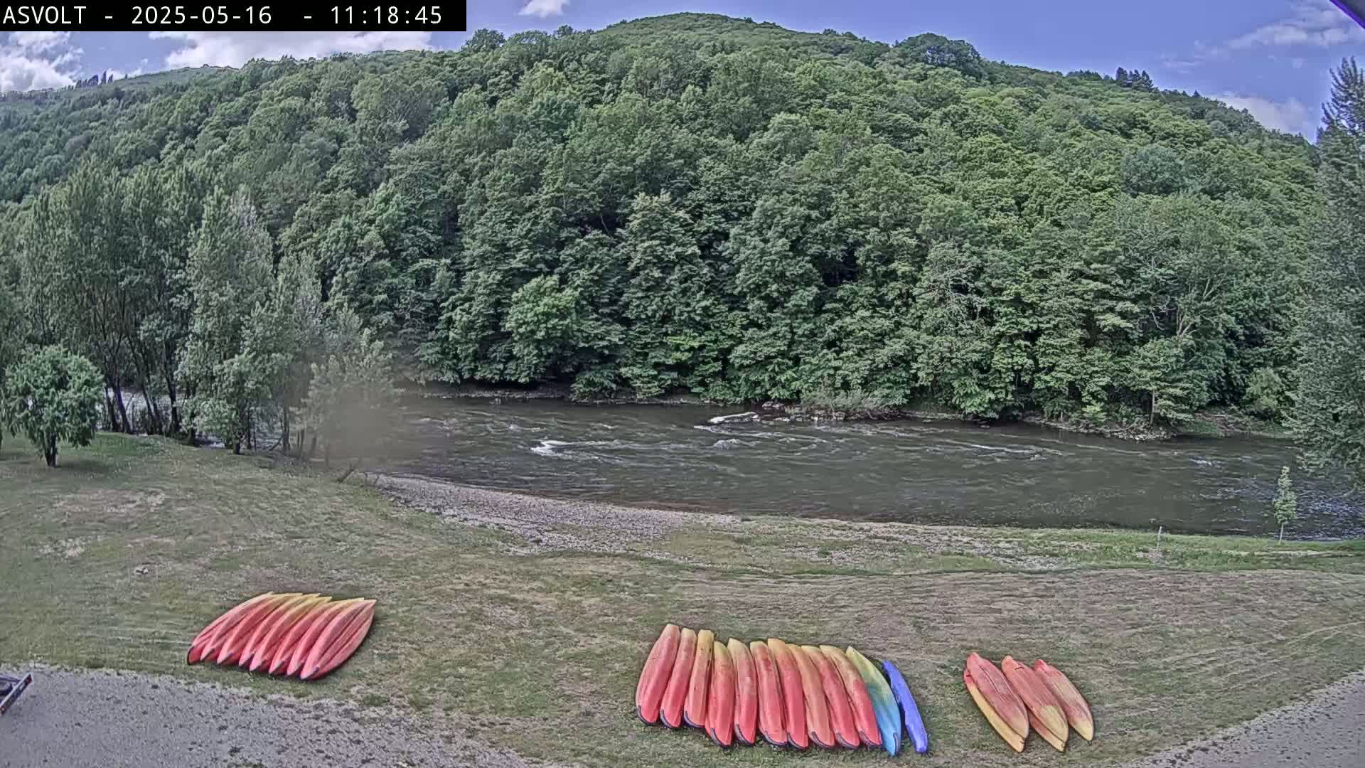 Kayaks are stacked on the grassy bank of a river under a partly cloudy sky, with lush green hills in the background.