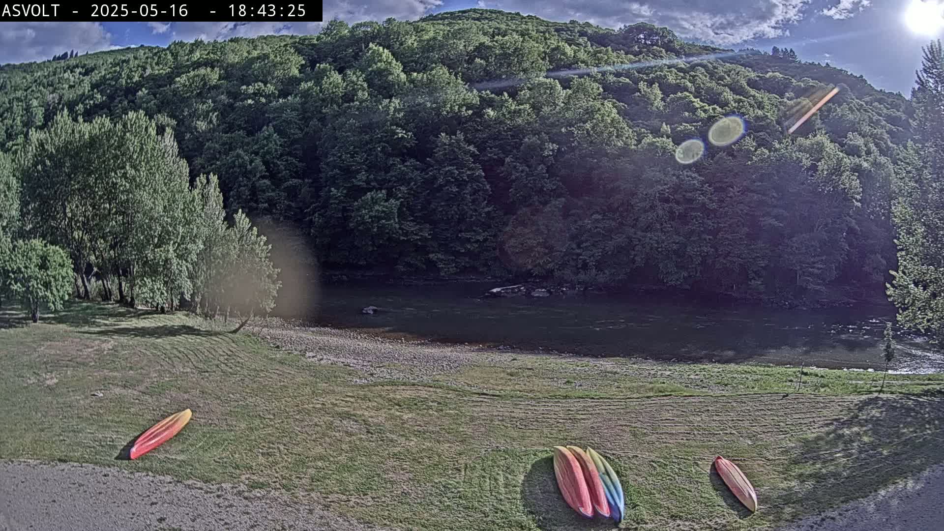 Several kayaks are lying on a grassy area next to a calm river under a partly sunny sky, with a lush green hillside in the background.