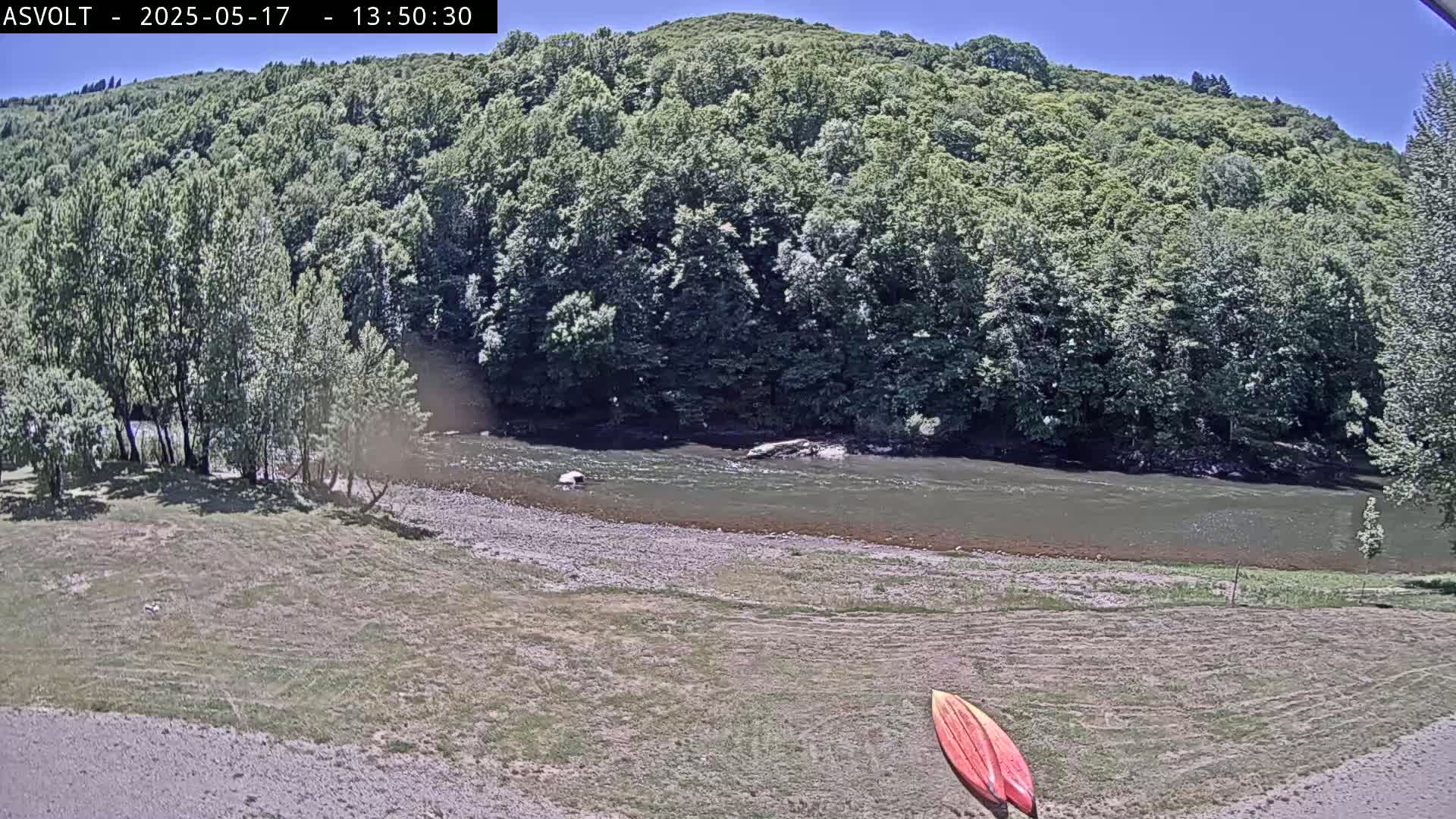 A river flows beneath a verdant hillside on a sunny day, with a pair of kayaks lying on the grassy riverbank.