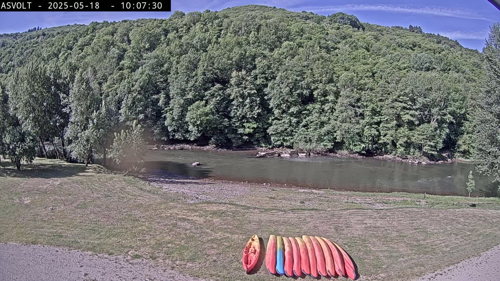 Several kayaks are lined up on a grassy bank next to a calm river under a mostly sunny sky, with lush green hills in the background.