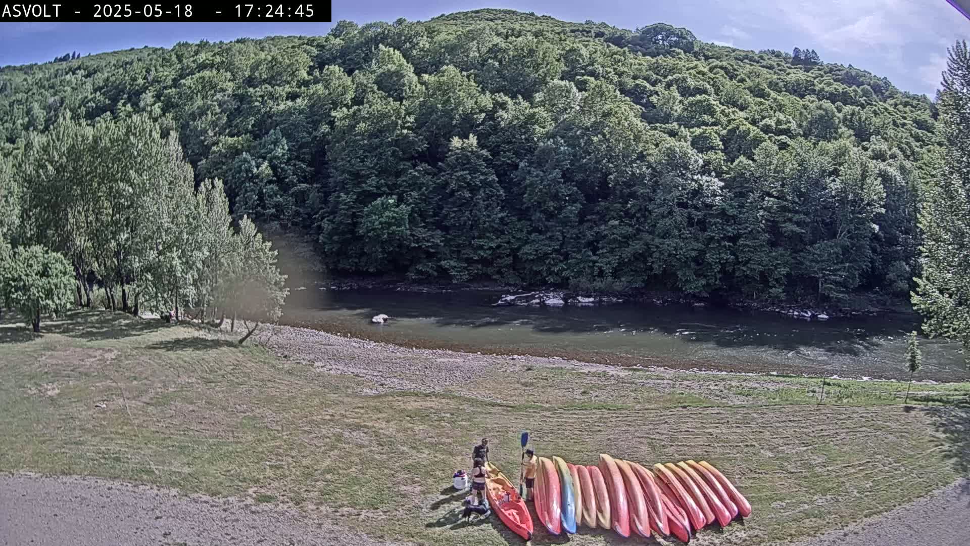 A grassy riverbank, under a mostly sunny sky, features several kayaks lined up near two people, with a calm river and lush green hills in the background.