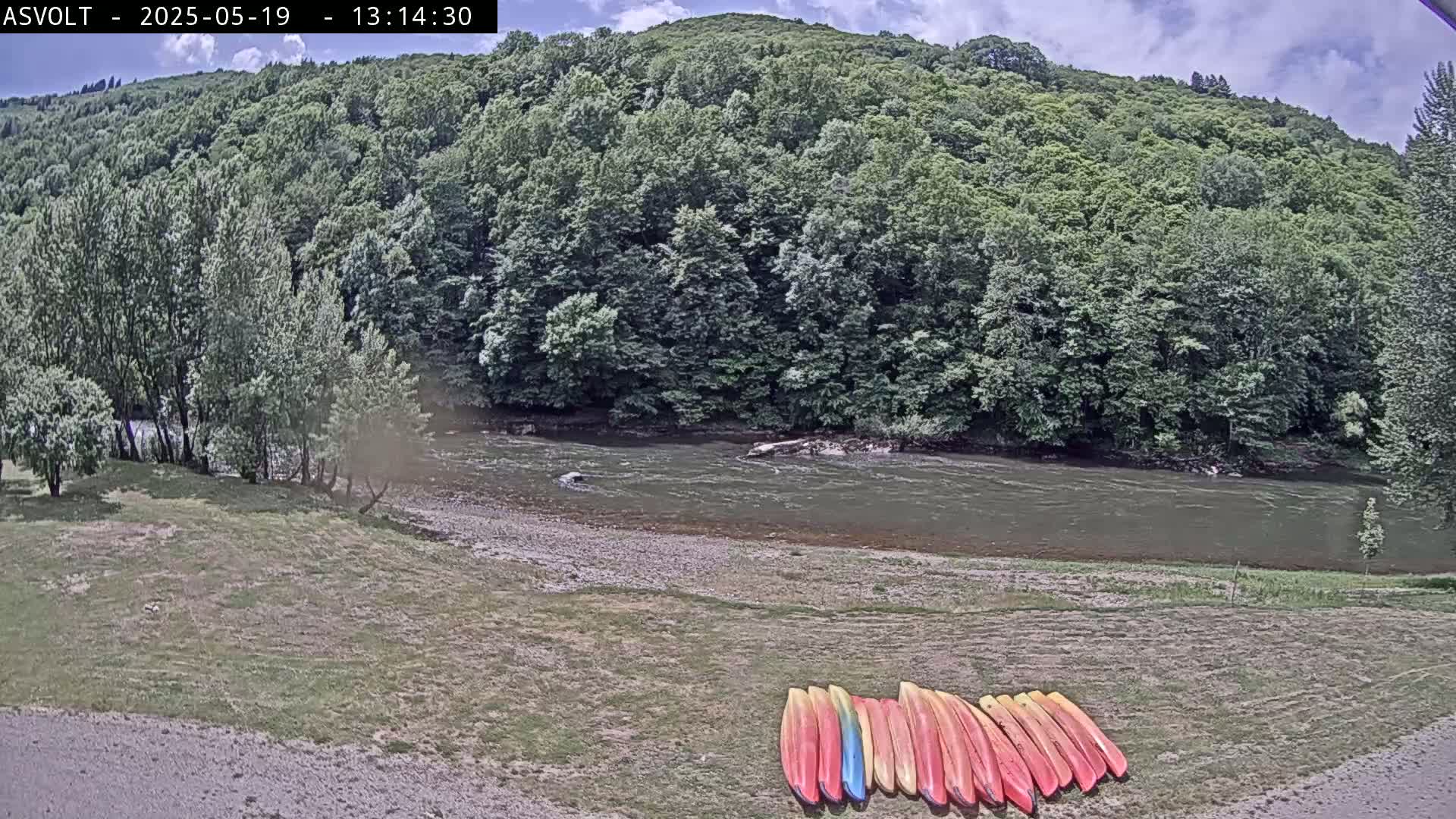 A partly cloudy day shows a river flowing past a lush green forest, with a group of kayaks lined up on the riverbank.