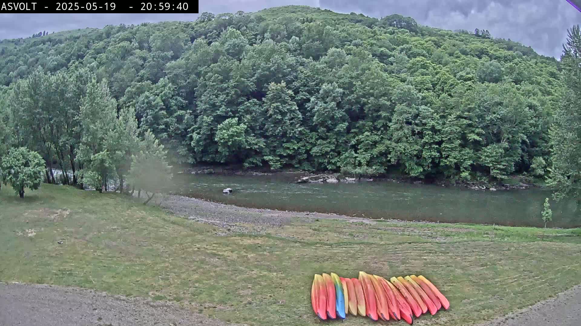 A calm river flows past a grassy area where several colorful kayaks are lined up, all under a cloudy sky and surrounded by lush green forests.