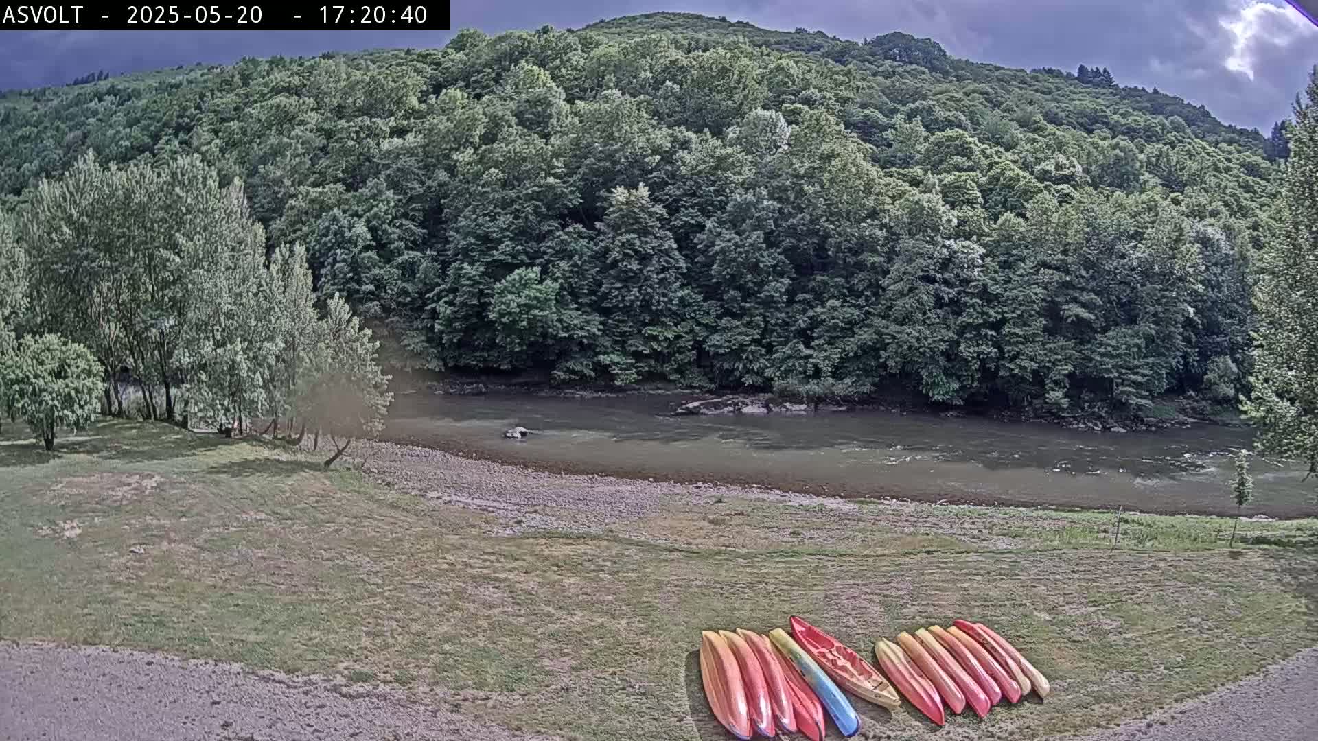Several kayaks are lined up on a grassy bank next to a calm river that flows beside a lush green forest under a partly cloudy sky.
