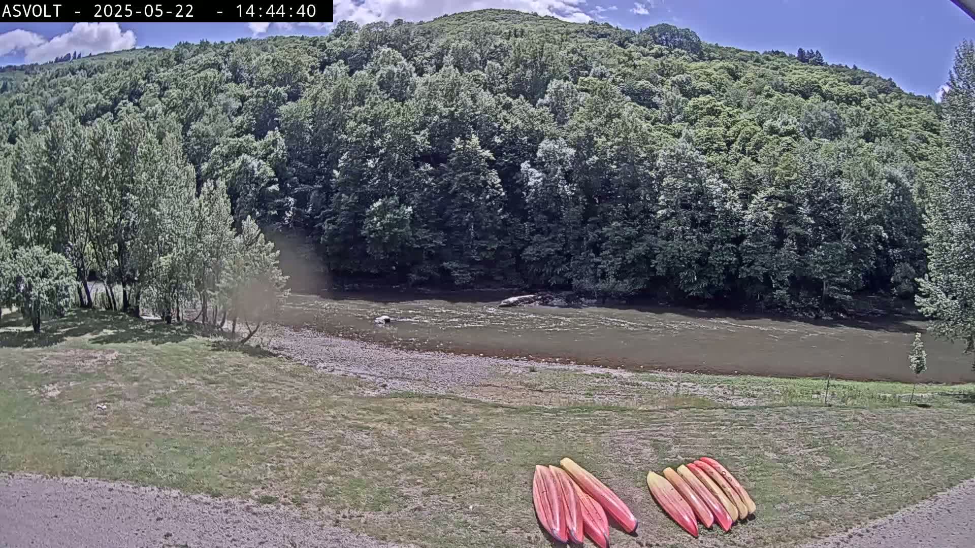 Several red and yellow kayaks are lined up on a grassy bank next to a calm, brown river flowing past lush green trees under a partly sunny sky.