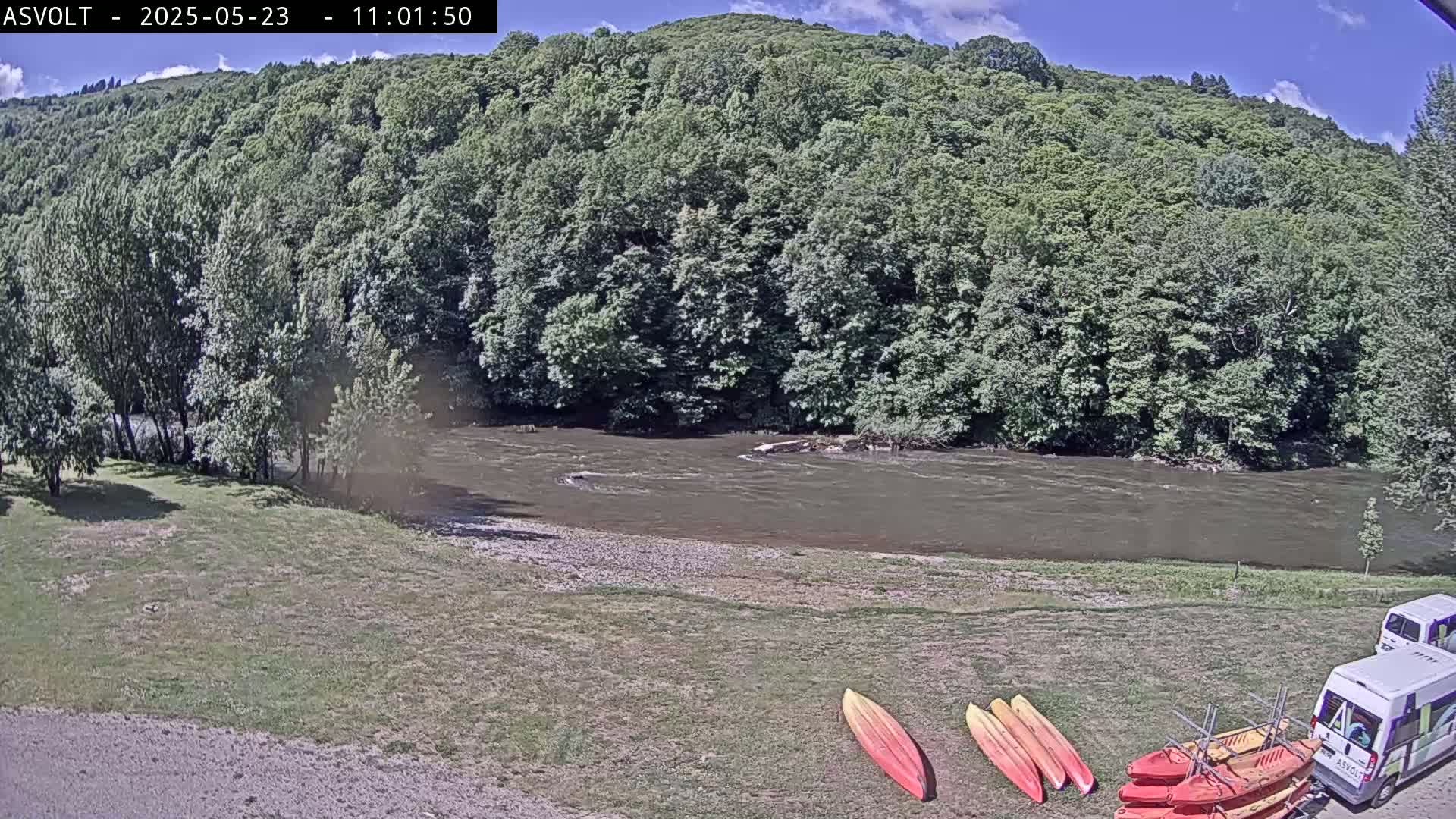 Kayaks are lined up on the grassy bank of a murky river under a partly sunny sky, with lush green hills in the background.