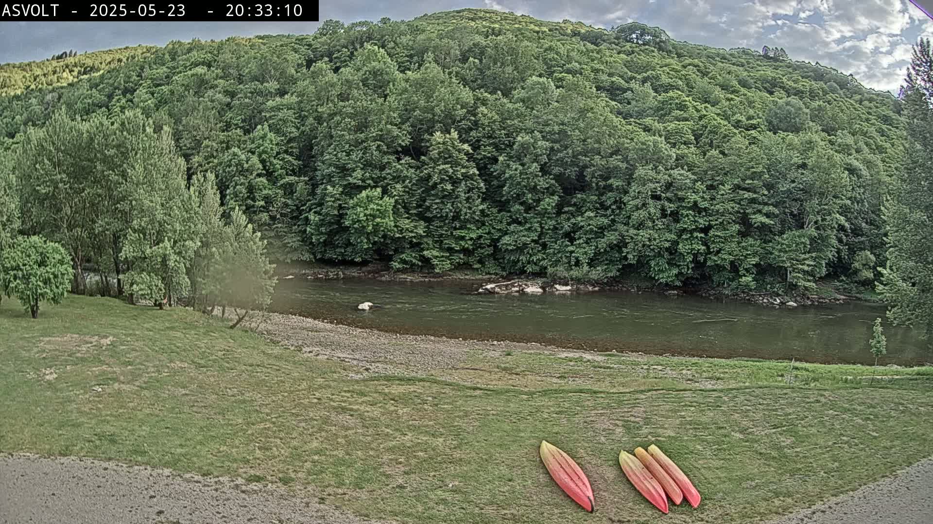 Three kayaks lay on a grassy area next to a calm river bordered by lush green trees under a partly cloudy sky.