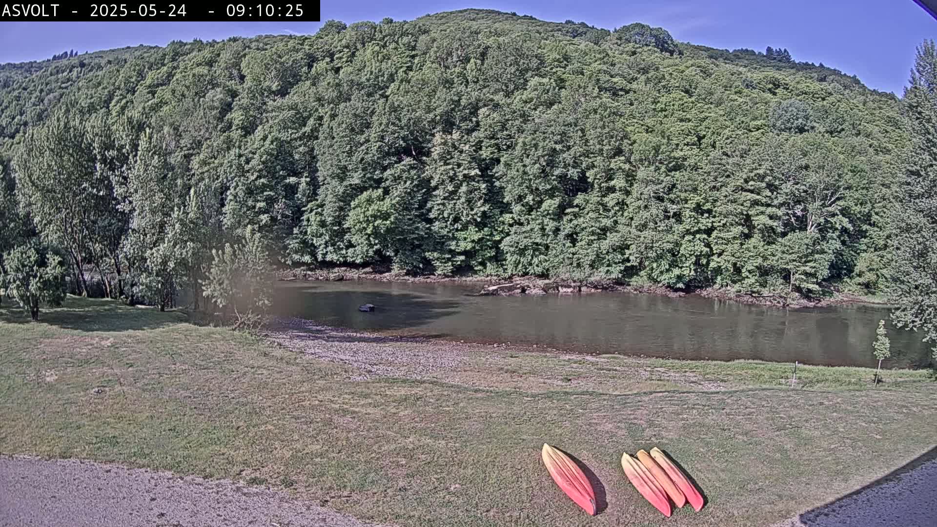 Three kayaks lay on a grassy area next to a calm river, which is bordered by lush green trees and a hill under a clear, sunny sky.