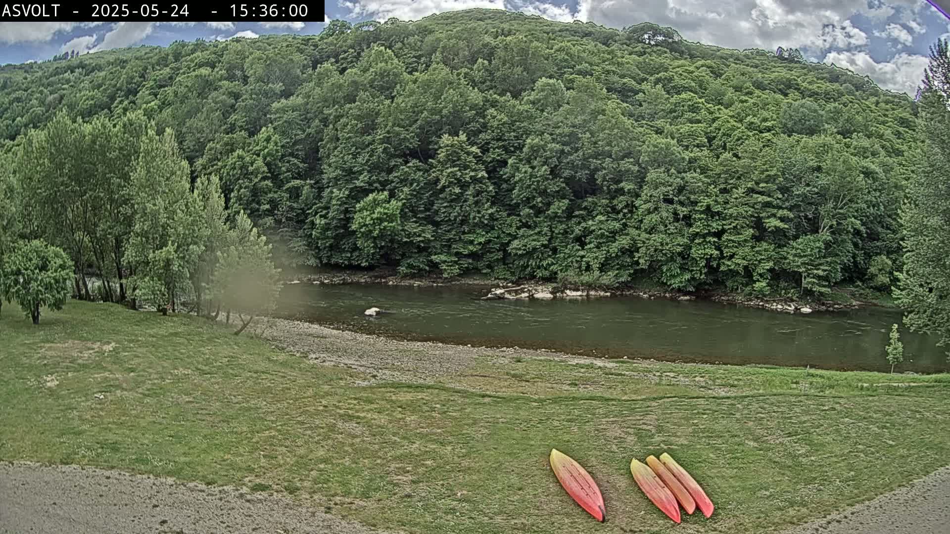Three kayaks lie on a grassy area beside a calm river, backed by lush green forest under a partly cloudy sky.
