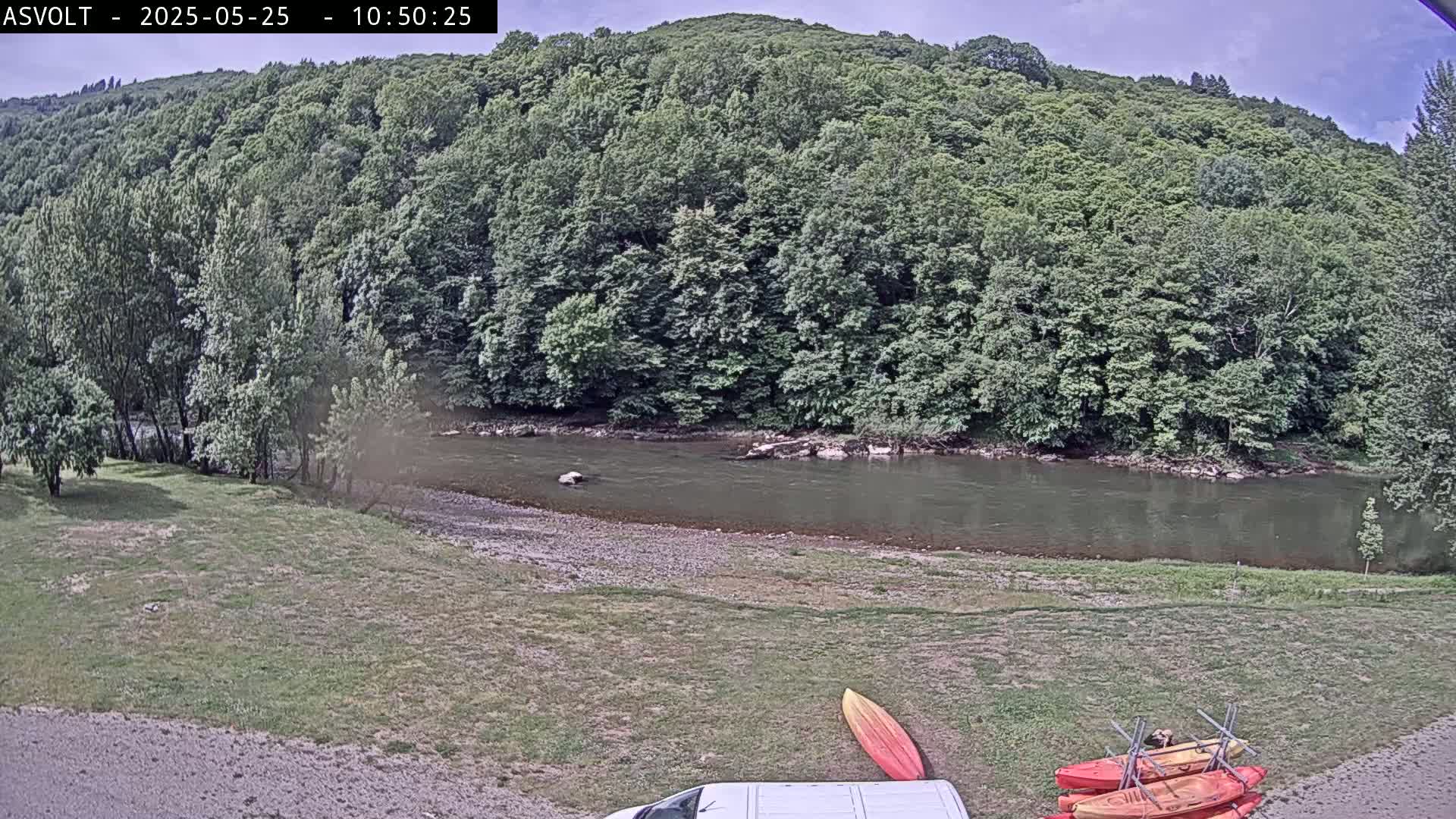 A calm river flows beside a grassy area with kayaks and a van parked nearby, all surrounded by lush green trees under an overcast sky.