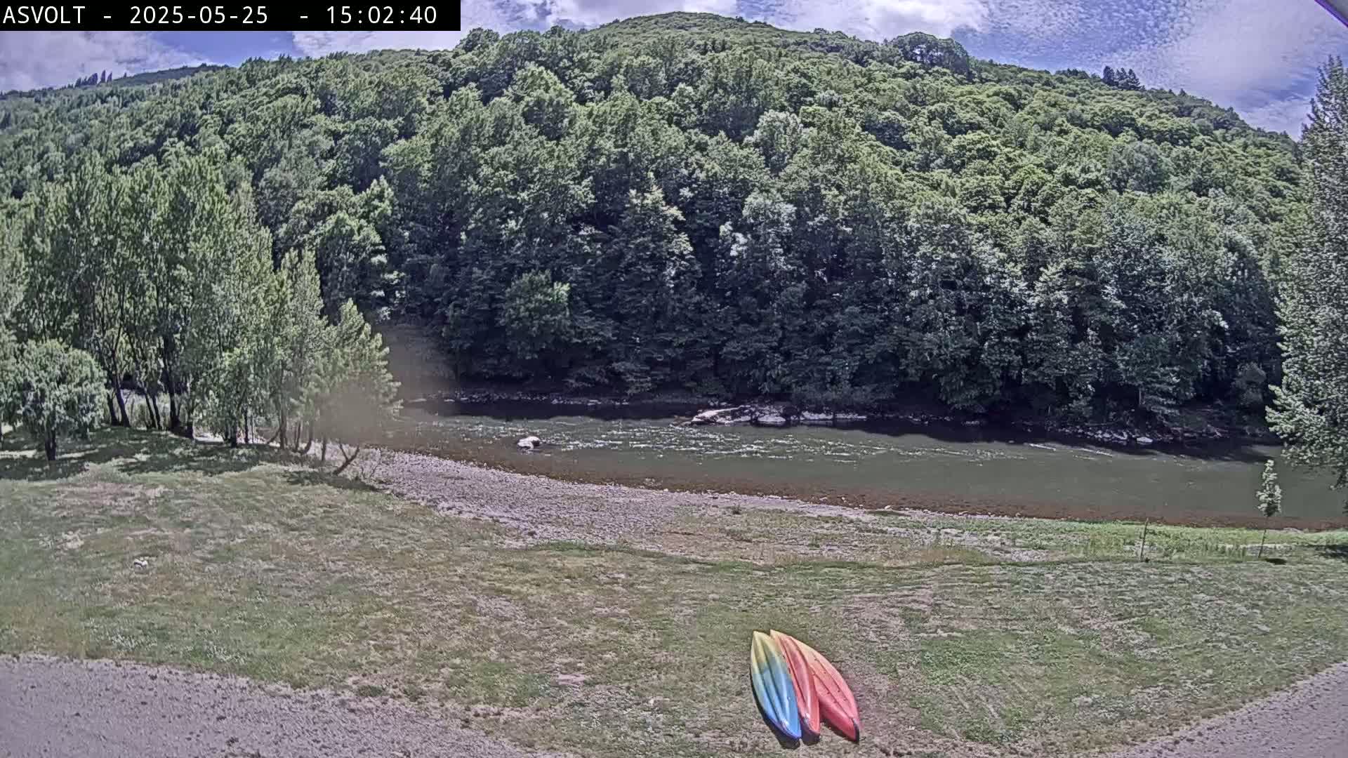 Two kayaks lie on a grassy bank beside a calm river flowing through a lush green forest under a partly cloudy sky.