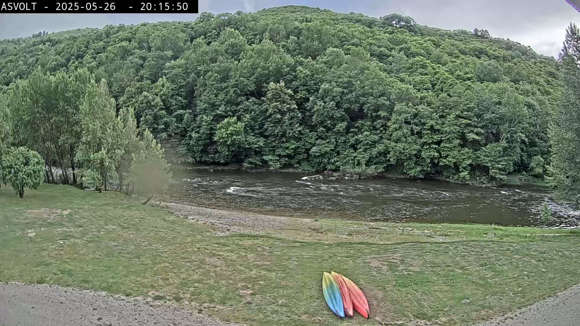 Two kayaks lay on a grassy area next to a flowing river bordered by lush green trees under a cloudy sky.