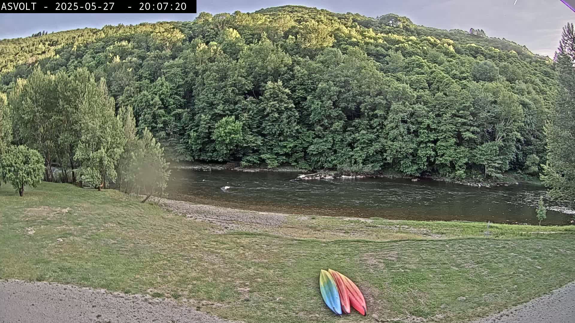 Two kayaks lie on a grassy area beside a calm river that flows past a lush green hillside under a partly cloudy evening sky.