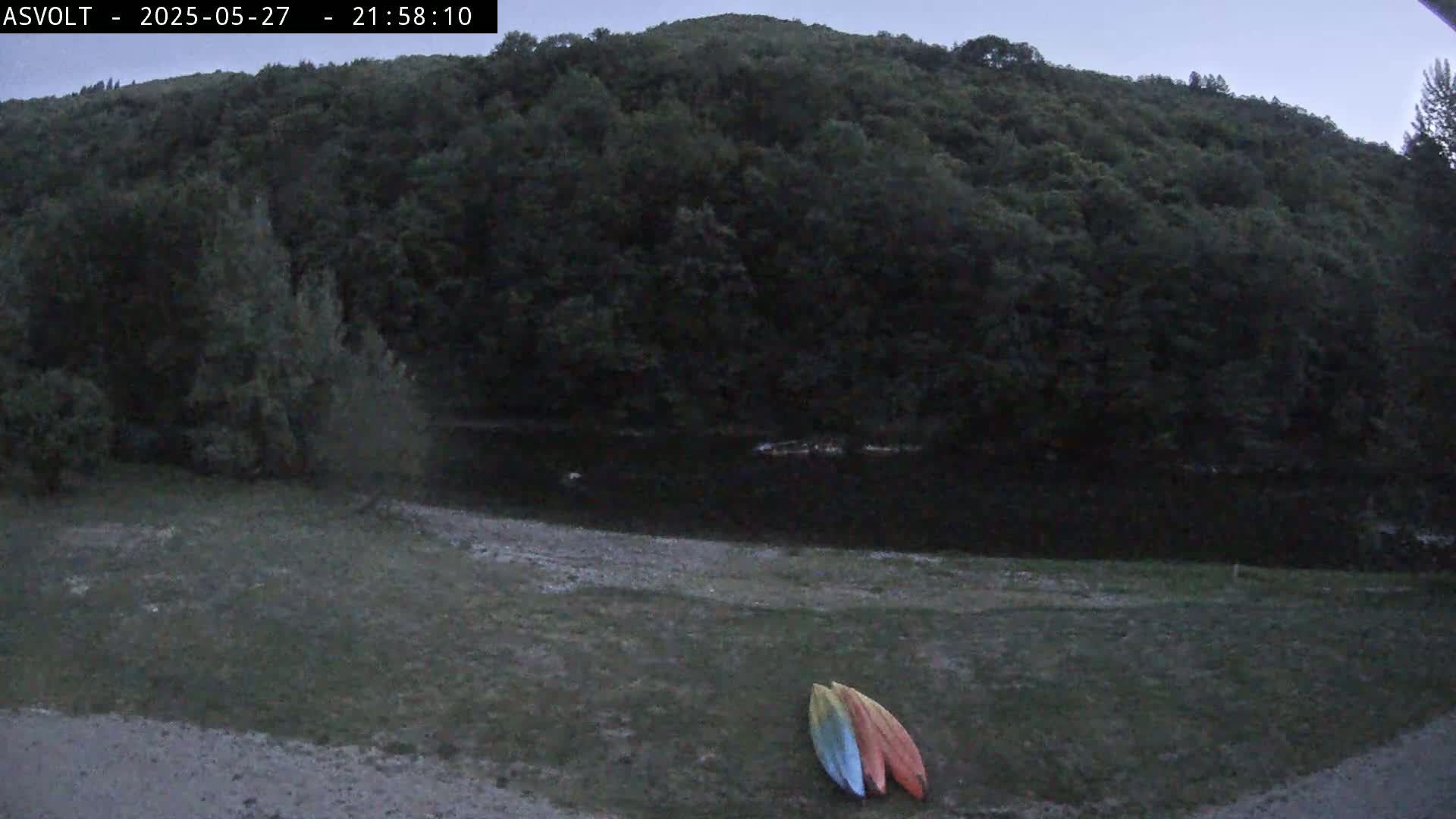 Two kayaks lie on the grass near a calm, dark river, backed by a heavily wooded hill under a twilight sky.