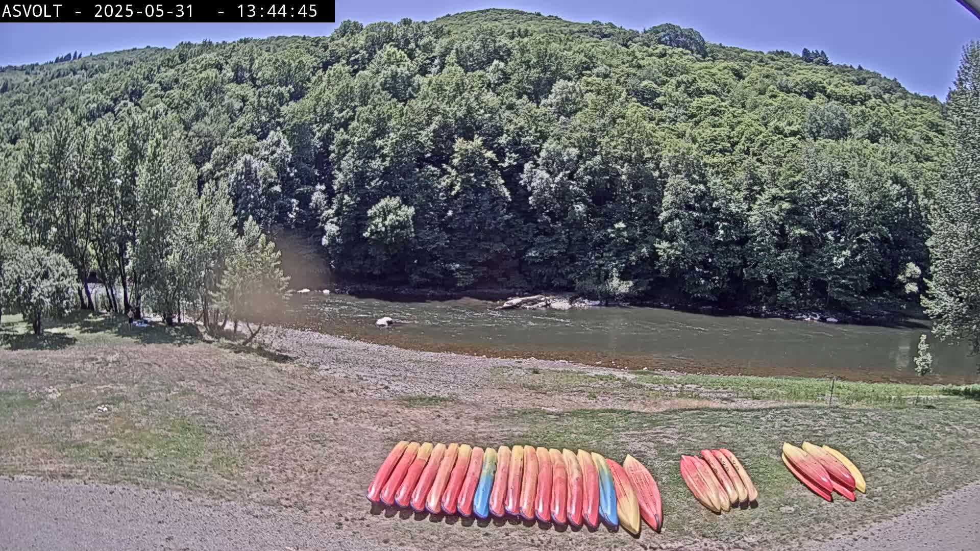 A calm river flows past a lush green forest, under a sunny sky; several colorful kayaks are lined up on the riverbank.