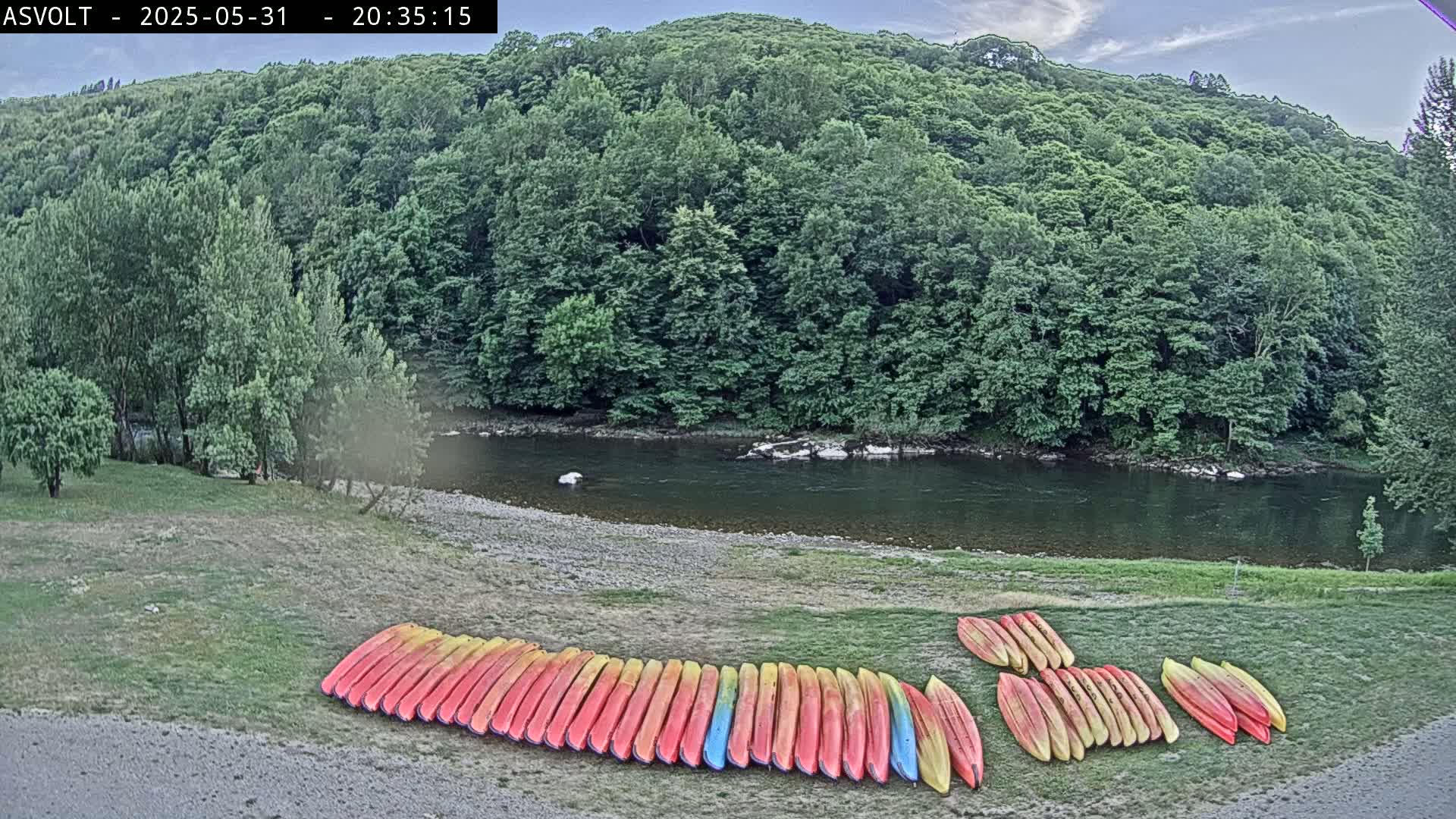 Numerous kayaks are lined up on a grassy bank next to a calm river under a partly cloudy sky, with a lush green forest in the background.
