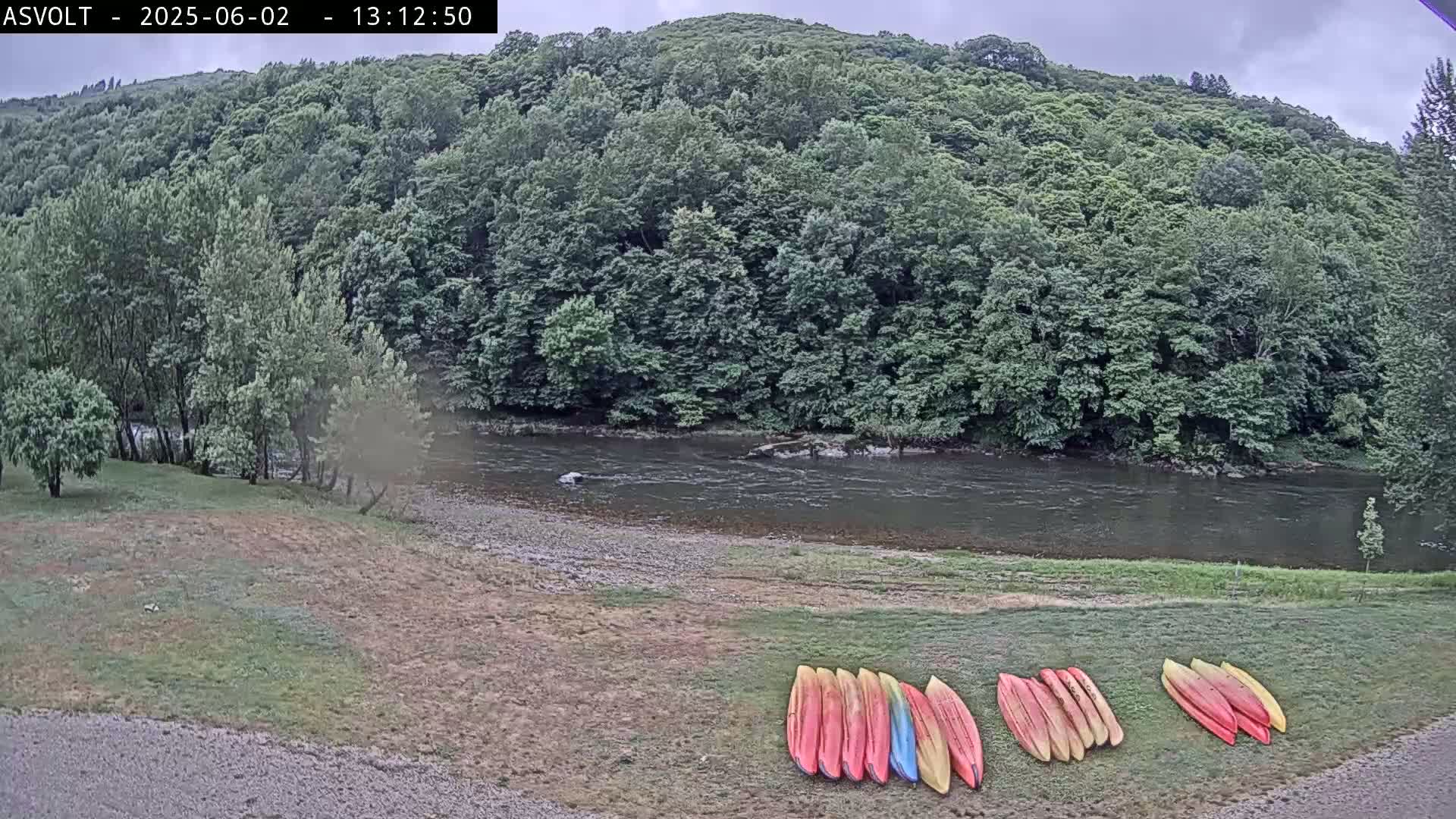 A calm river flows past a lush green forest under an overcast sky, with several kayaks lined up on the grassy riverbank.