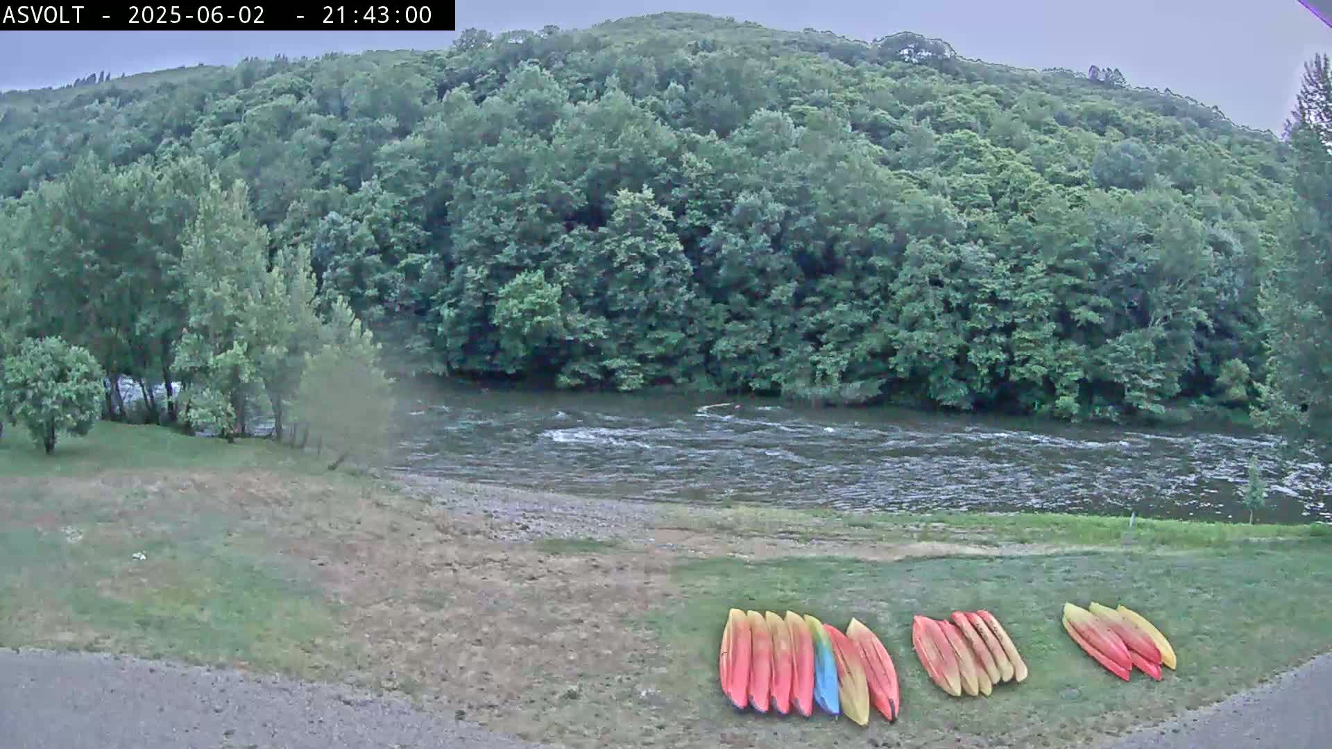 Several kayaks are lined up on a grassy area next to a flowing river, which is bordered by lush green trees under an overcast sky.