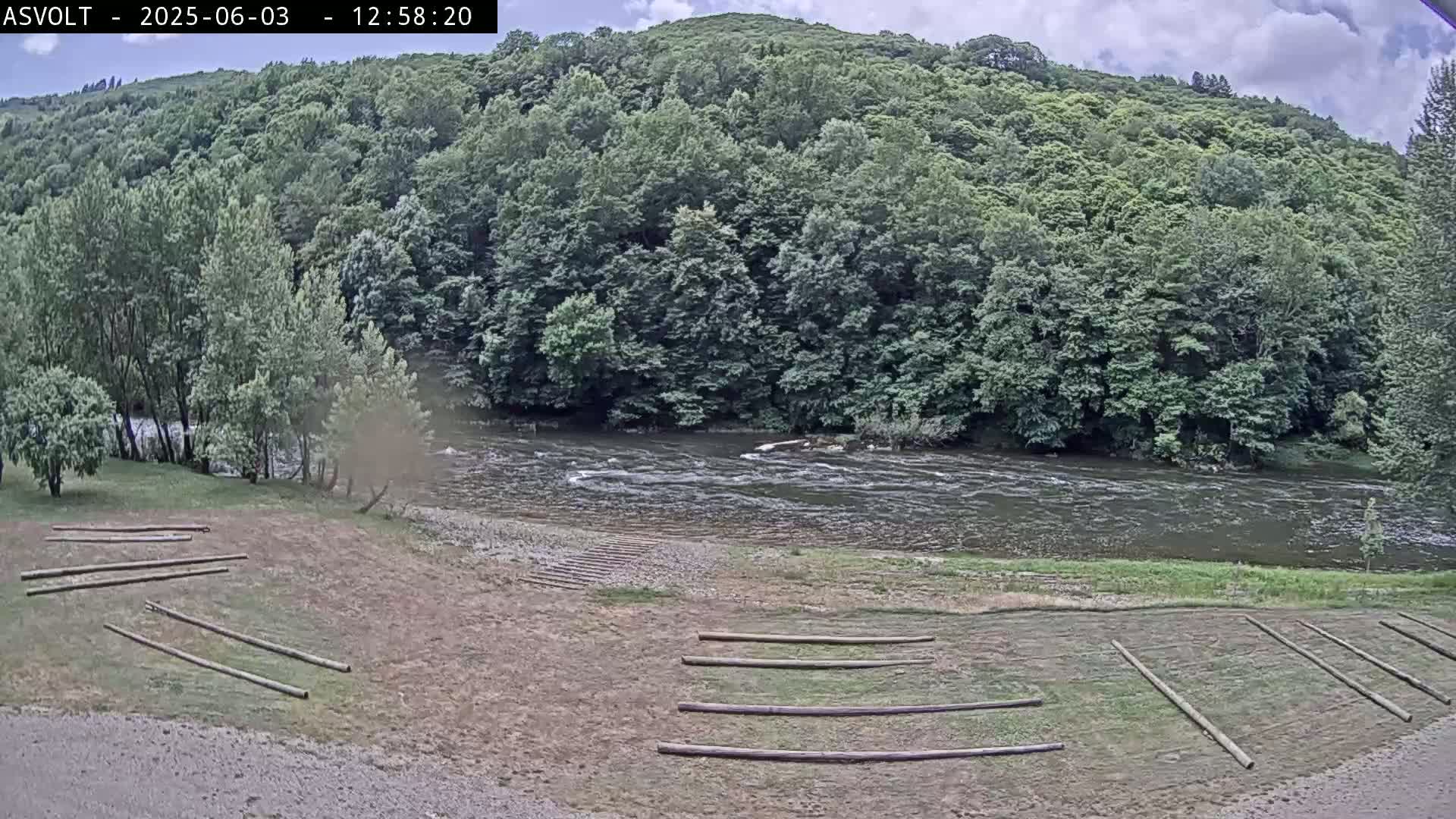 A partly cloudy day reveals a river flowing beside a grassy area with several wooden poles lying on the ground, all backed by a lush green forest covering a hillside.