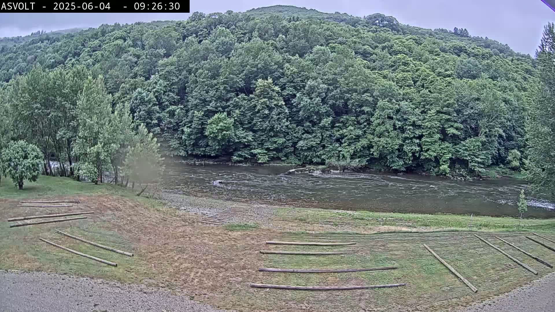 A grey, overcast day reveals a river flowing past a grassy area with stacked lumber, bordered by lush green trees and hills.