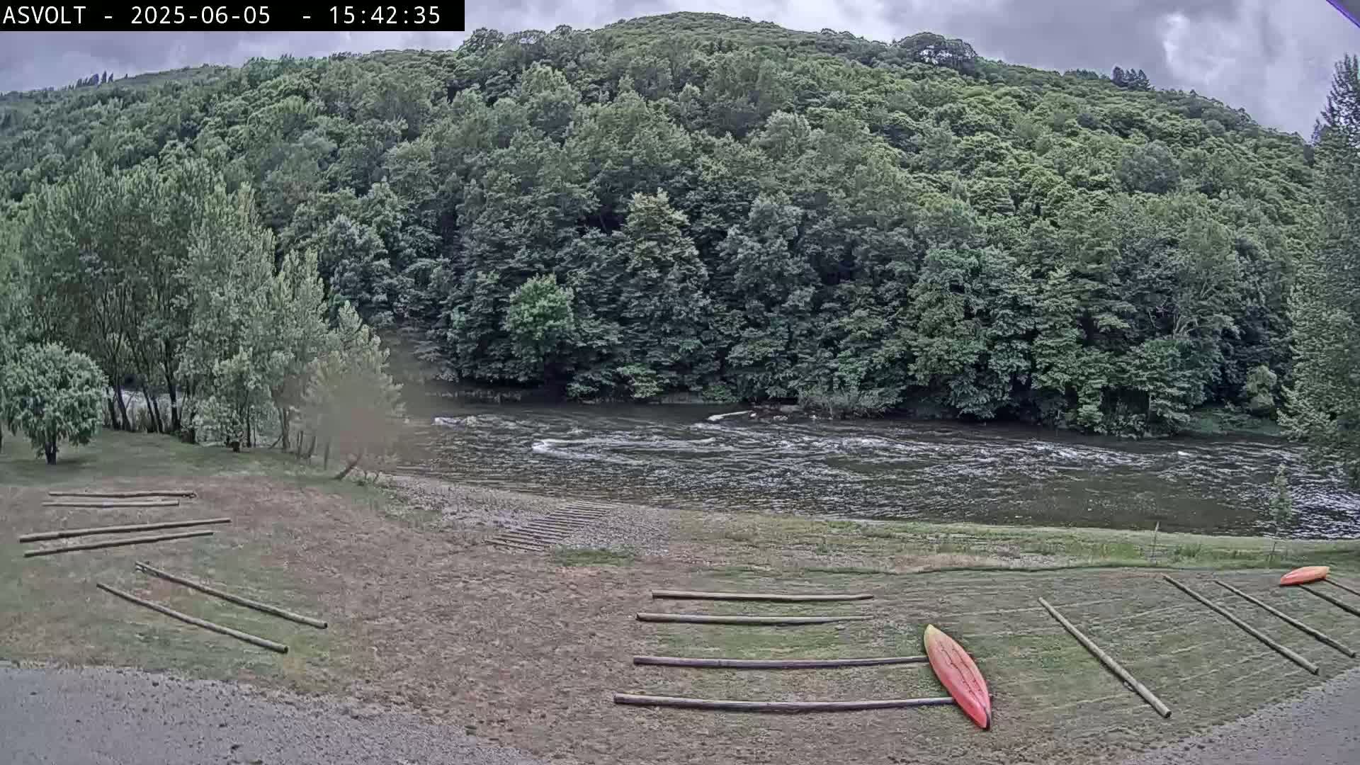 A river flows under a cloudy sky past a wooded hillside and a grassy bank where several kayaks are resting on wooden racks.