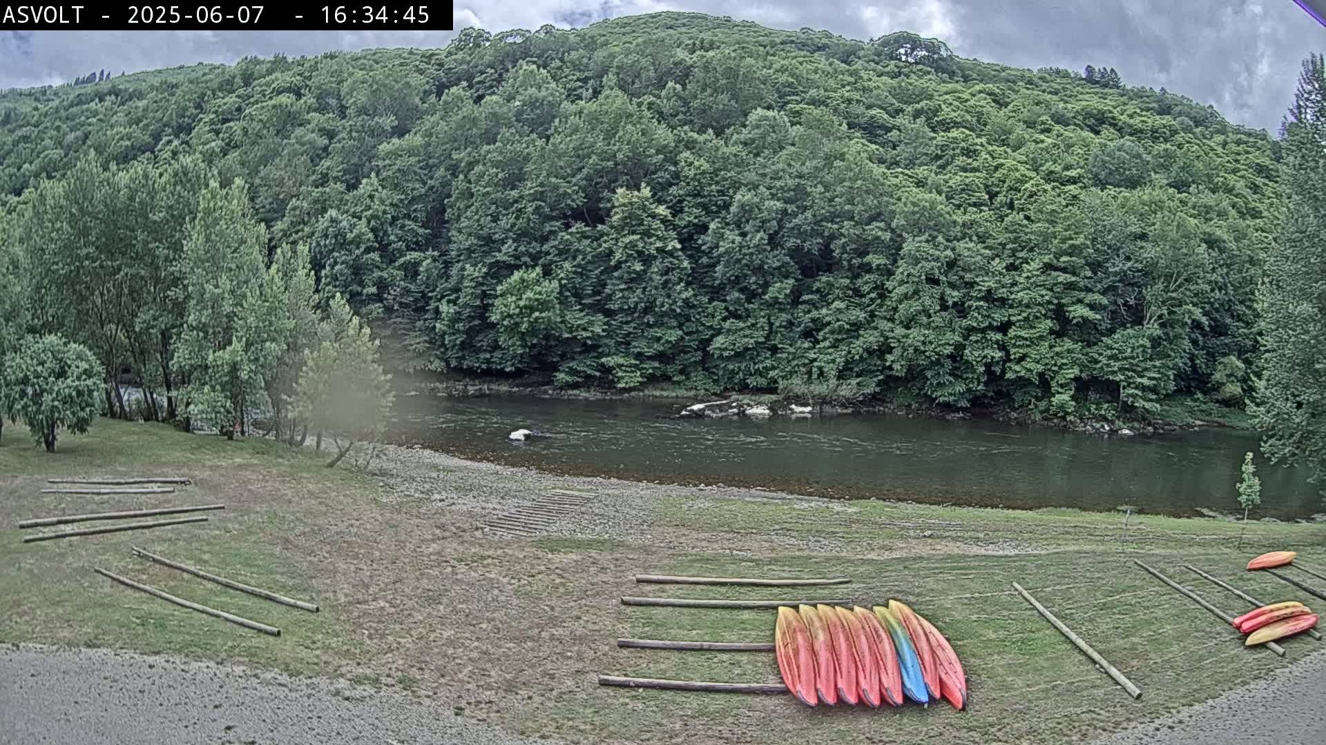 Kayaks are lined up on a grassy riverbank under a partly cloudy sky, next to a calm river flowing through a lush green forest.