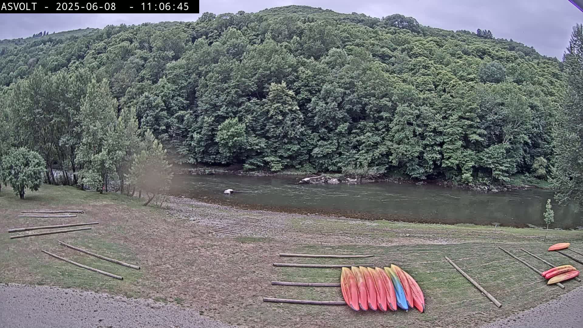 A river flows under an overcast sky past a wooded bank where several kayaks are stored on the grassy shore.