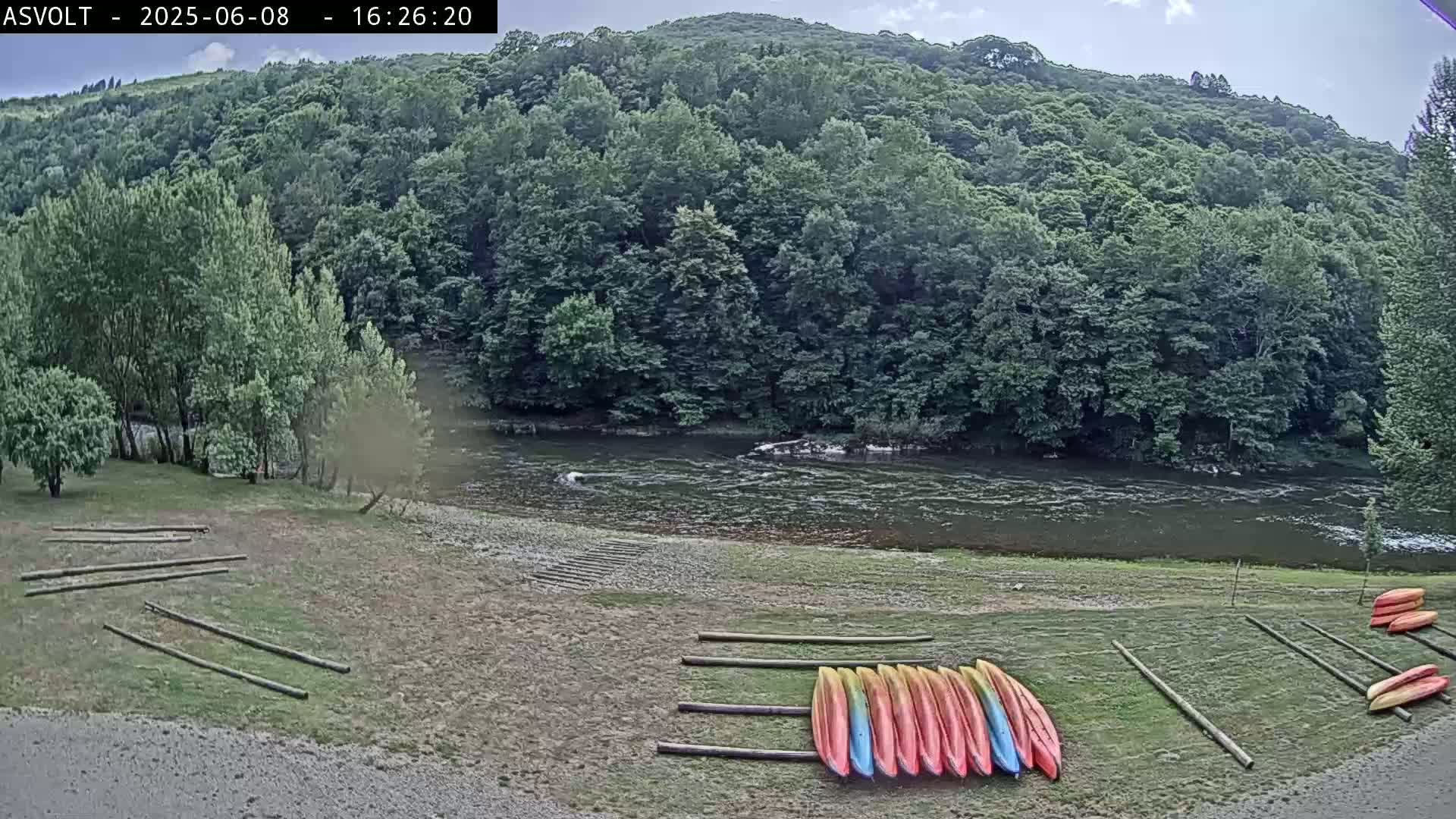 Kayaks are stored on racks near a river under a partly cloudy sky, with lush green hills rising in the background.