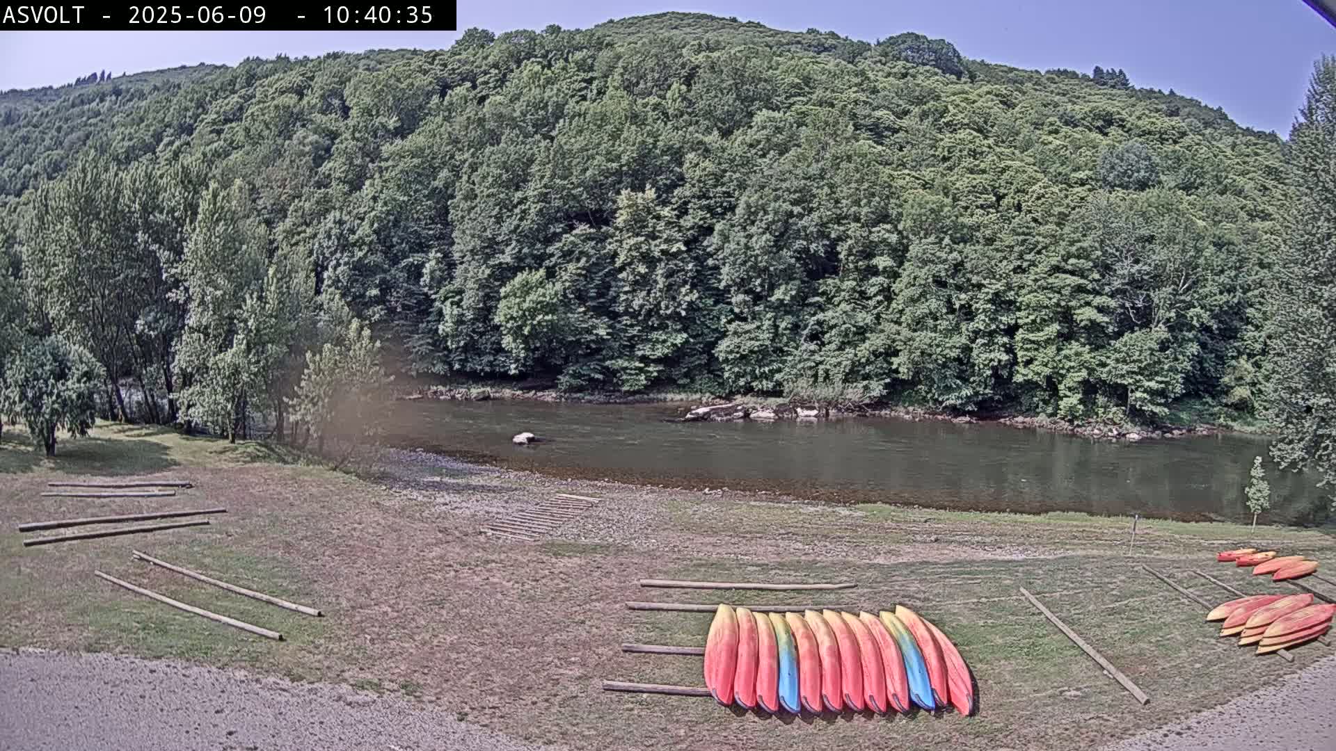 A calm river flows past a lush green forest, where numerous kayaks are lined up on a sandy riverbank under a clear, sunny sky.