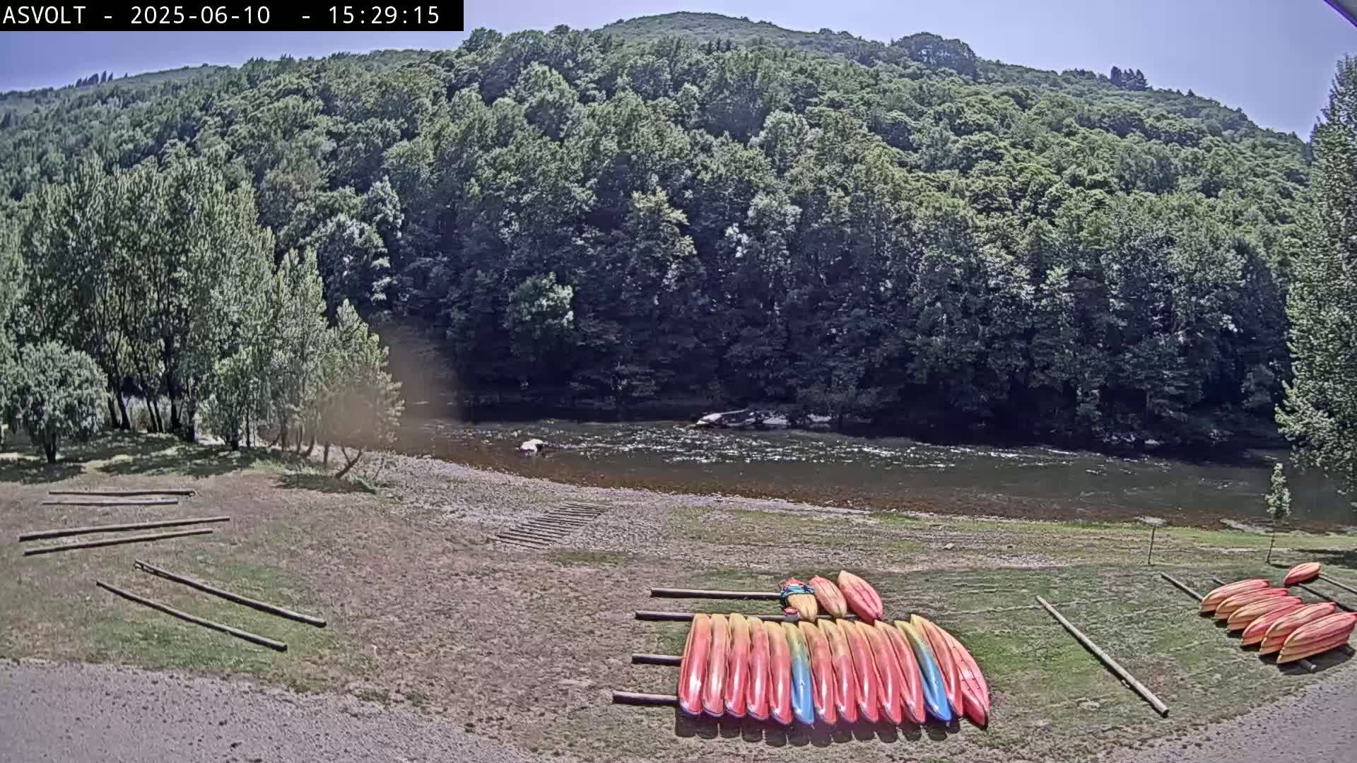 Kayaks are stored on racks next to a calm river under a sunny sky, with a lush green forest rising up behind it.