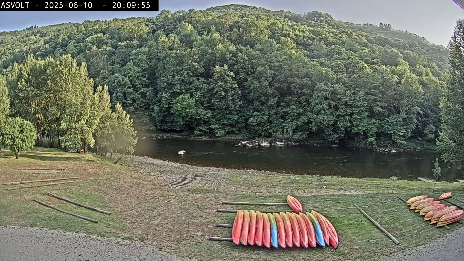 A calm river flows beneath a verdant hillside, where numerous kayaks are stored on the grassy riverbank under a partly cloudy sky.