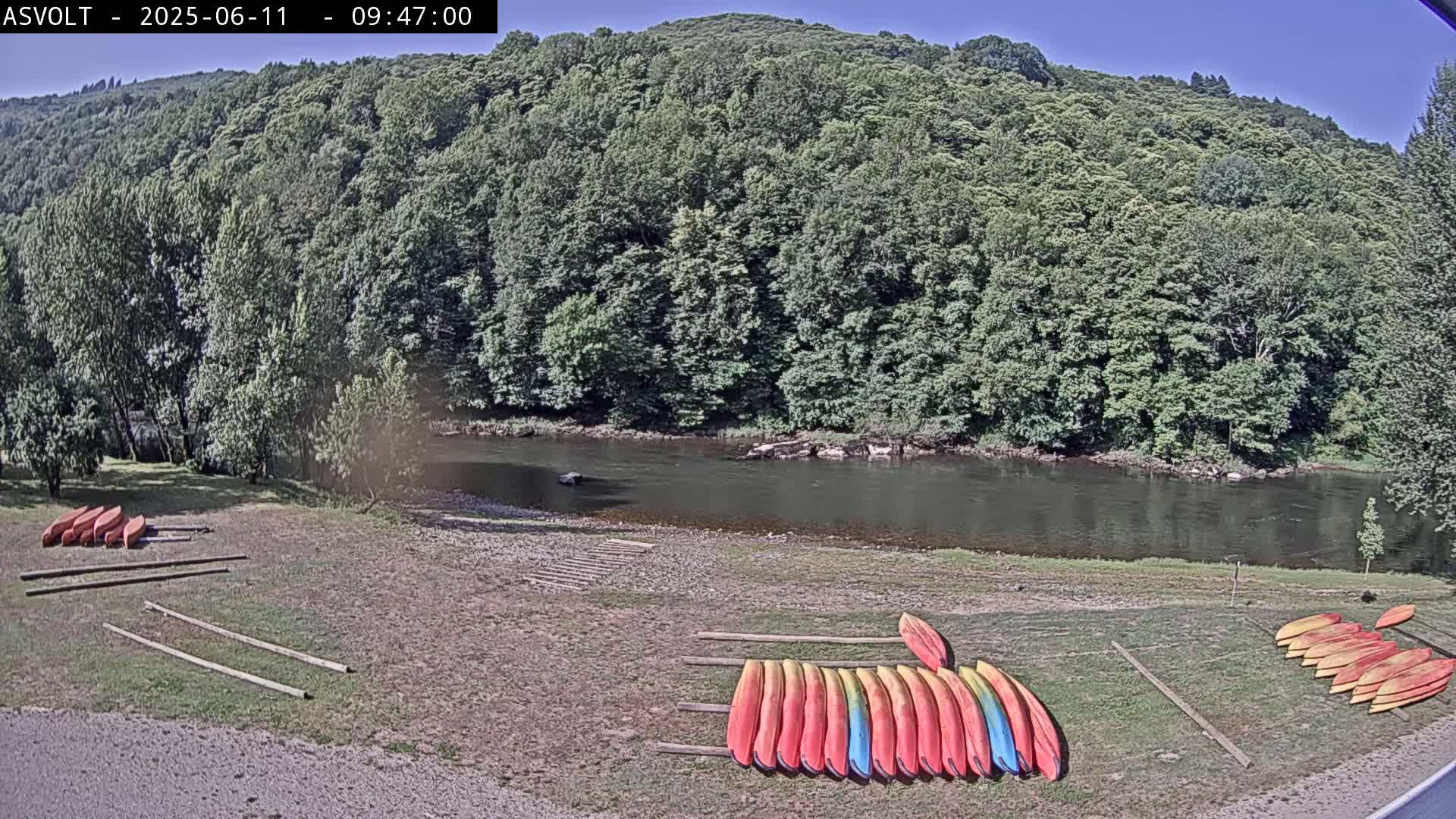 On a sunny day, numerous kayaks are stacked on the grassy bank of a calm river, next to a lush green forest.