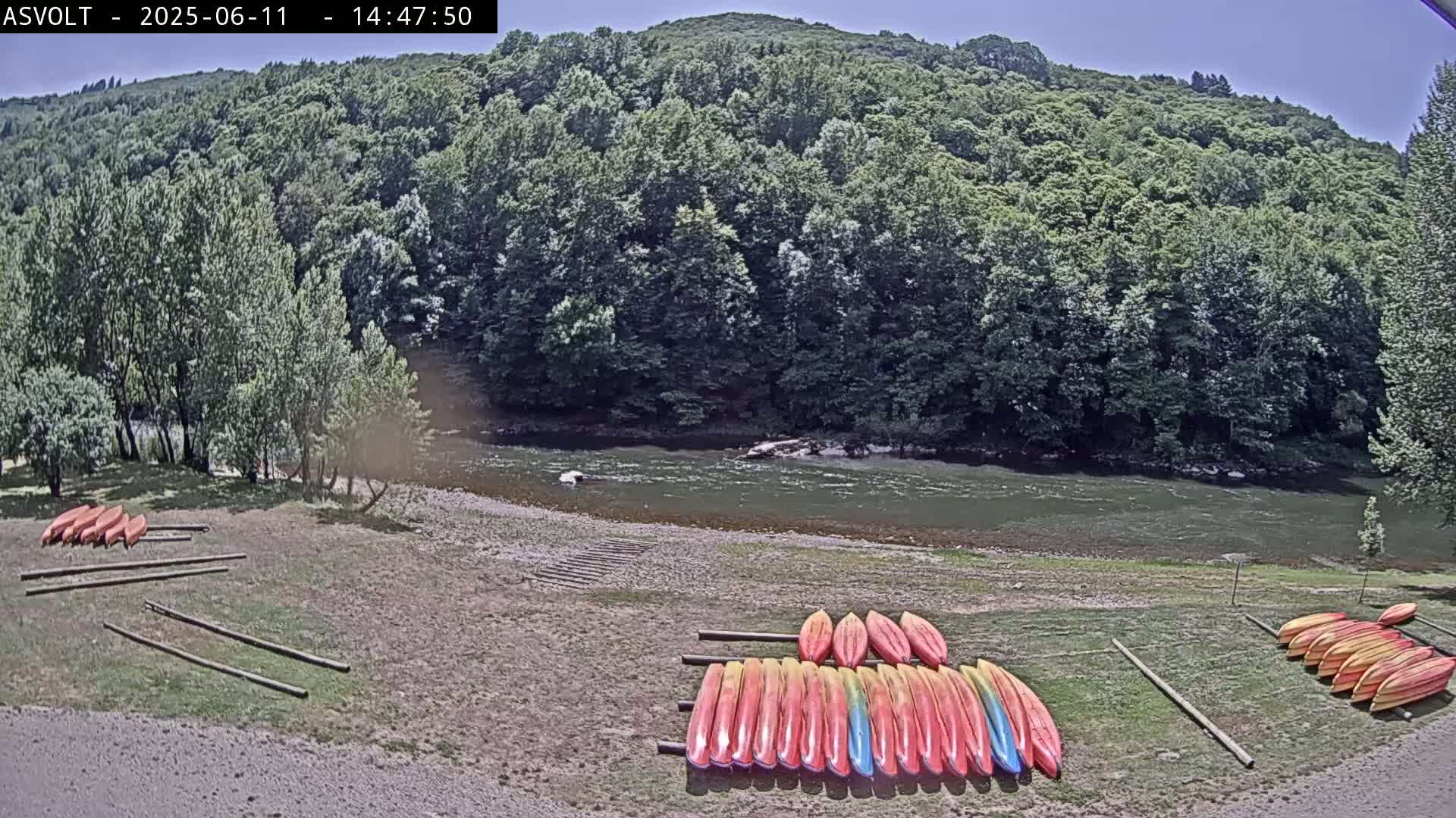 Kayaks are stacked on racks near a calm river, backed by a lush green forest under a sunny sky.