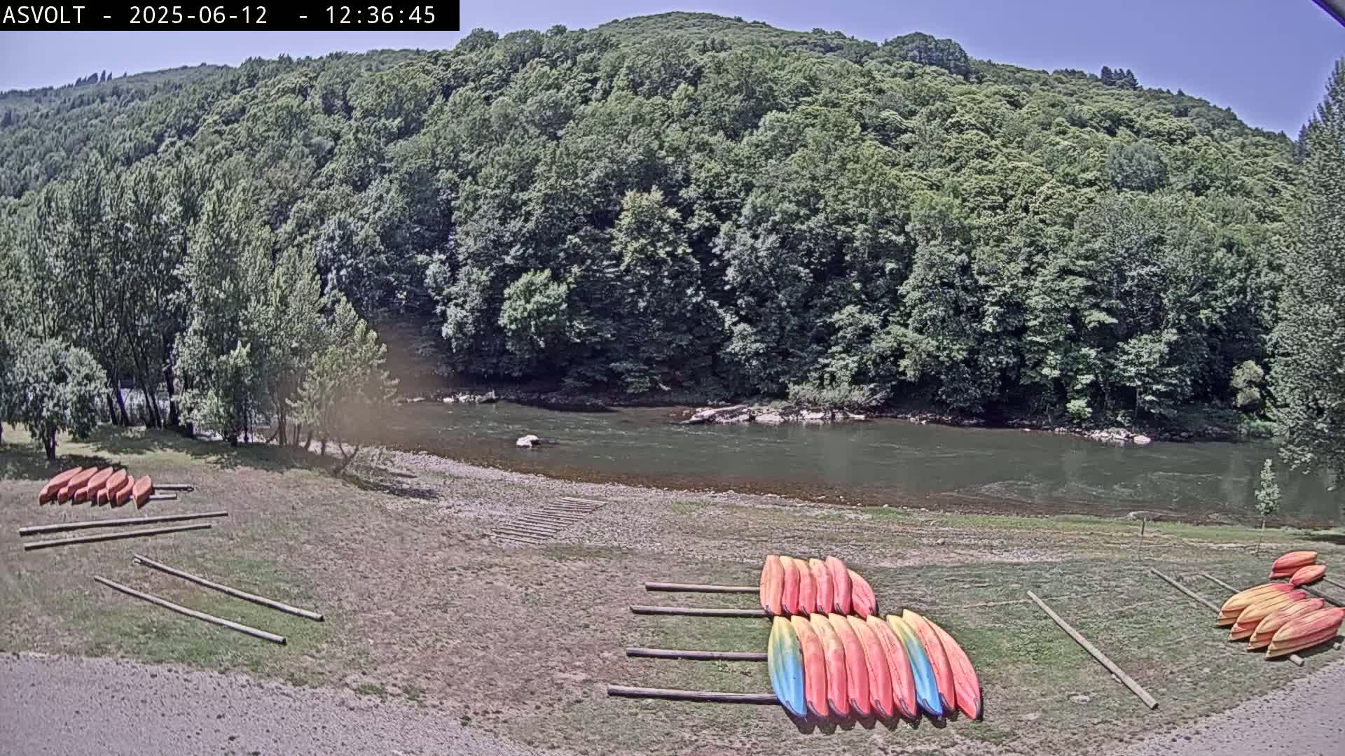 Kayaks are lined up on the shore of a calm river under a sunny sky, next to a lush green forest.