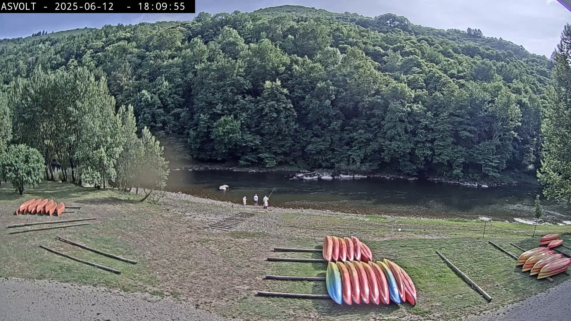 A calm river flows beneath a lush green hillside, where several kayaks are racked on the riverbank near three people fishing.