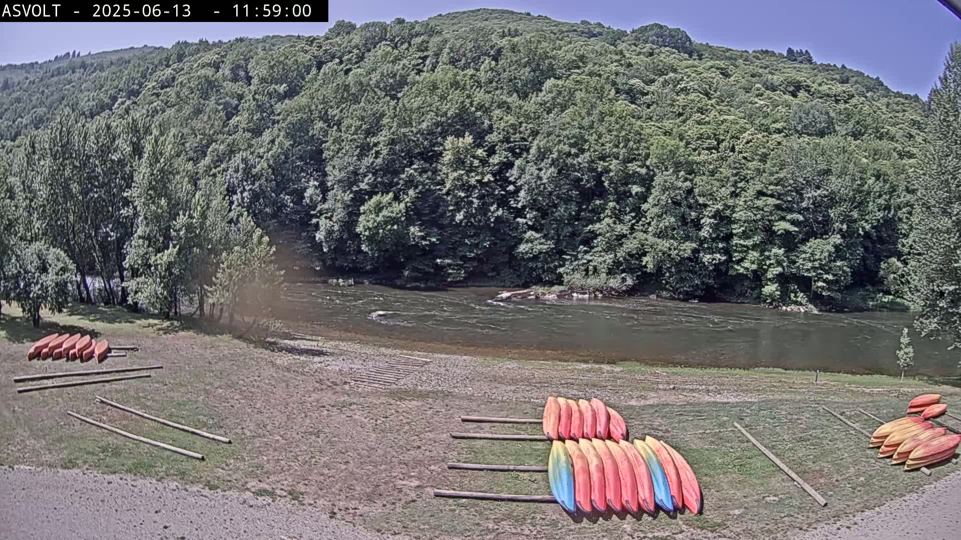 A sunny day shows a riverbank with numerous kayaks stored on racks, next to a calm river flowing past a lush green forest under a clear sky.