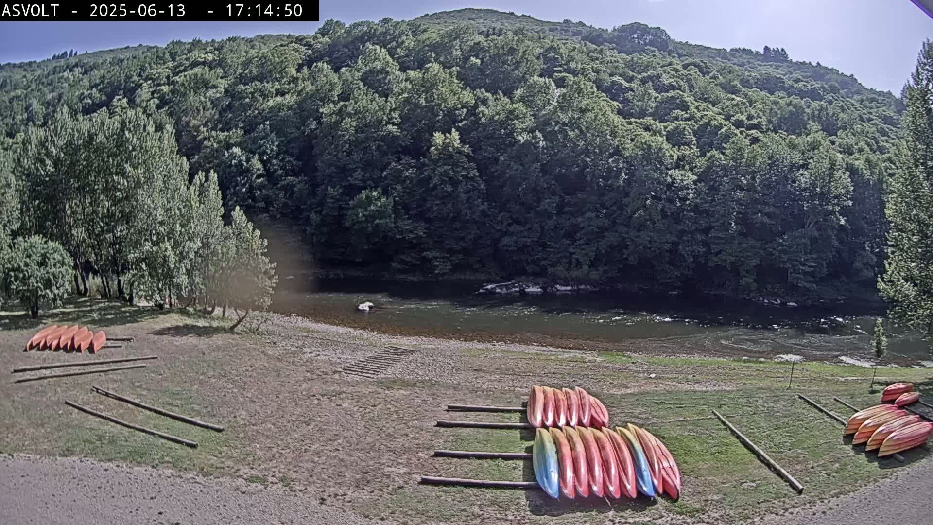 Several kayaks are stacked on the shore of a calm river, under a sunny sky, with lush green trees and hills in the background.