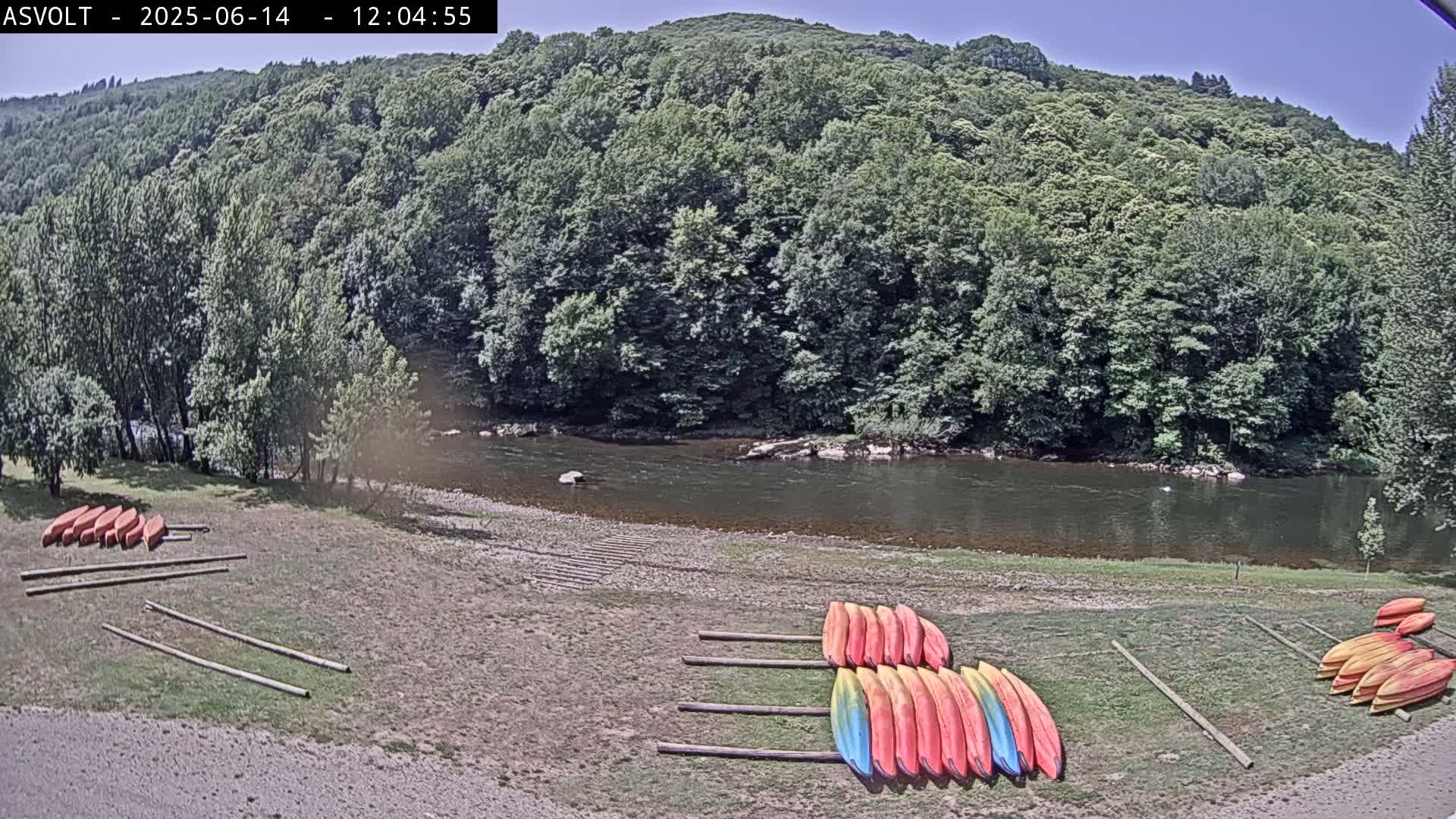 Under a clear sky, several kayaks are stored on wooden racks near a calm river, bordered by lush green trees and a hillside.
