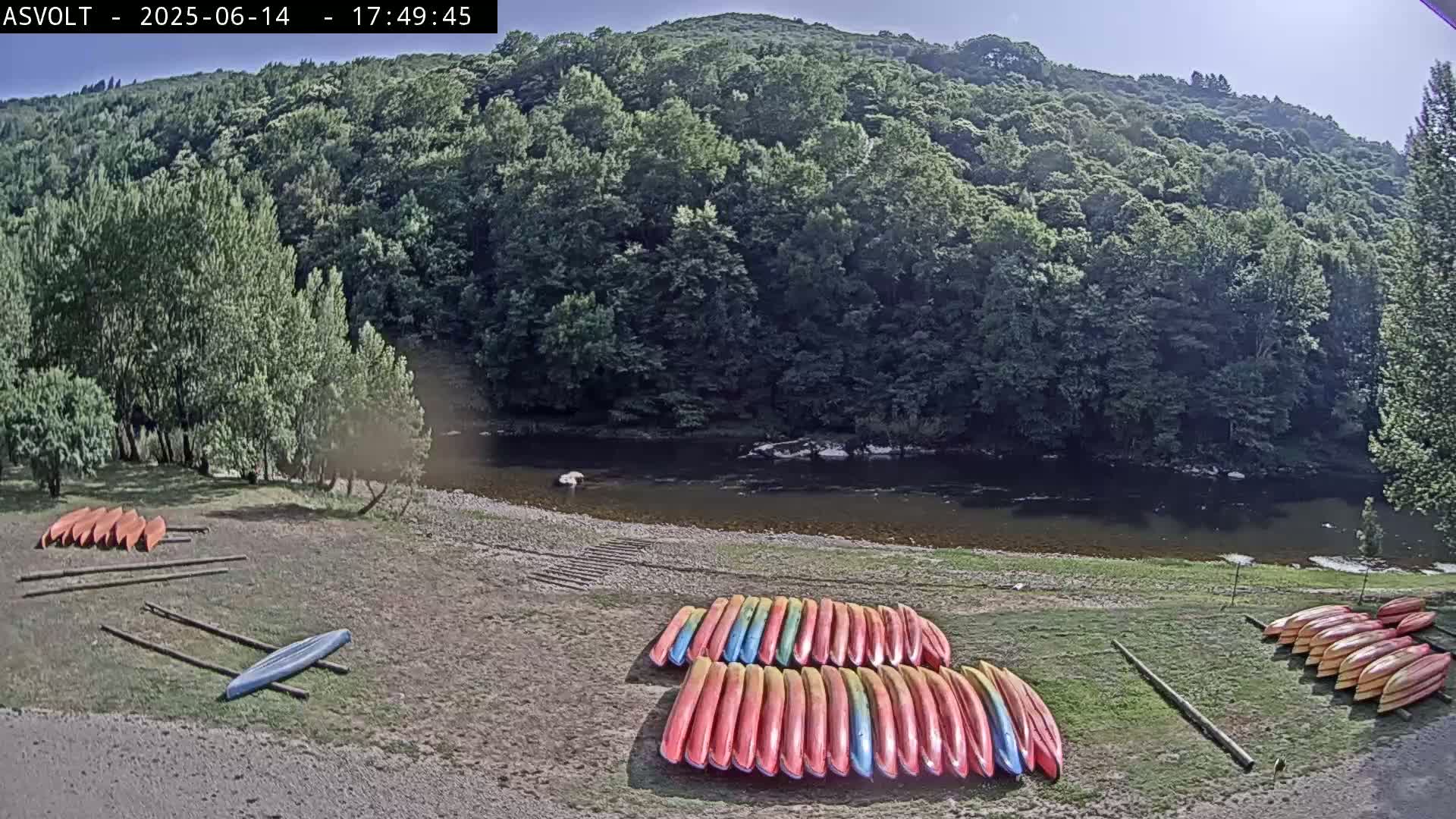 On a sunny day, numerous kayaks are stacked on the grassy bank of a calm river, bordered by lush green trees and hills.
